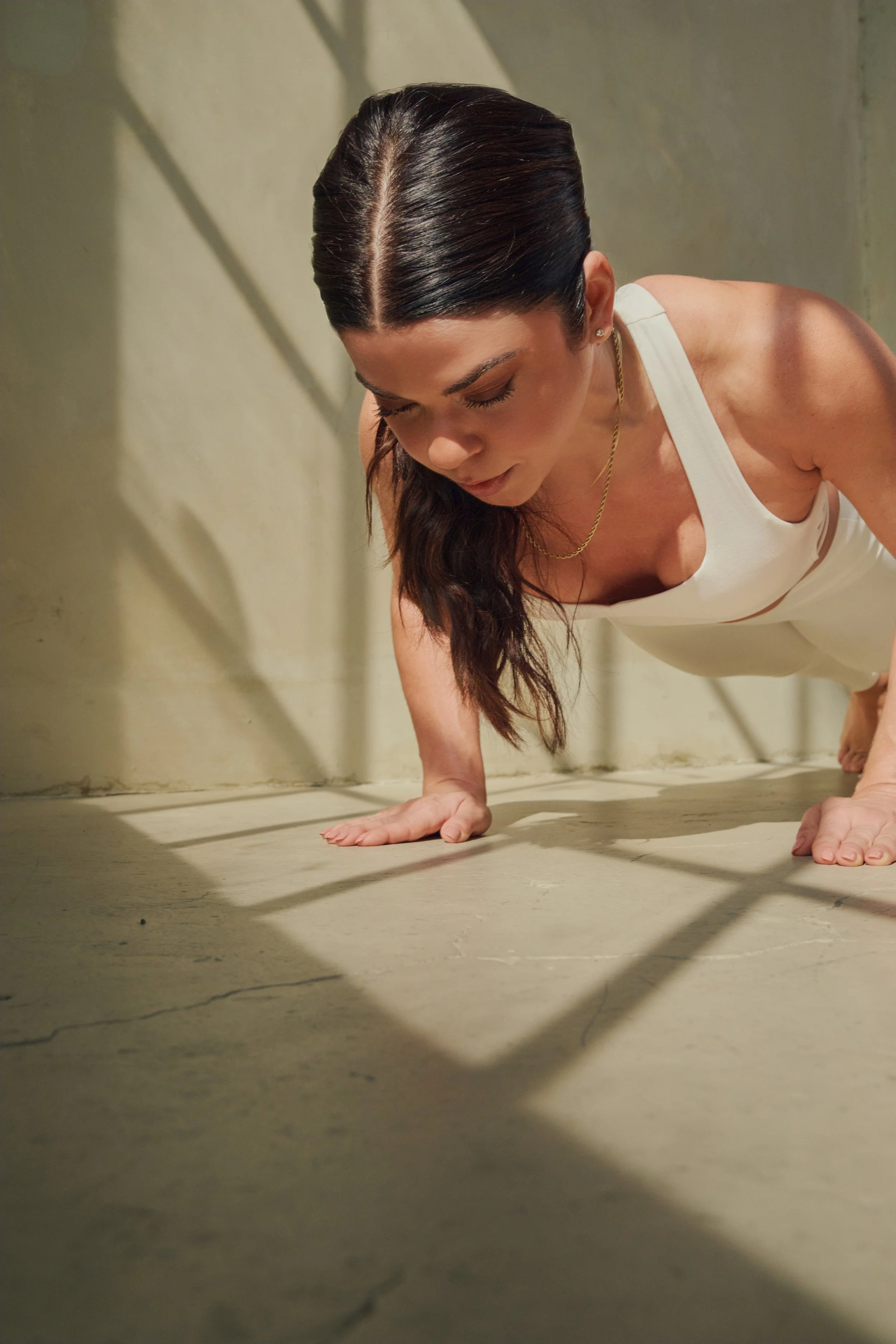 Woman in white tank top doing a push-up on the floor, with shadows cast on the wall and floor.