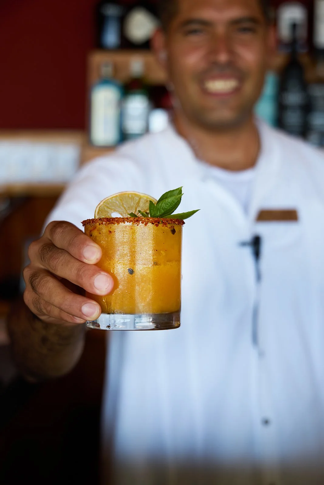 A bartender holding a yellow cocktail garnished with a lemon slice and basil leaves, smiling in the background.