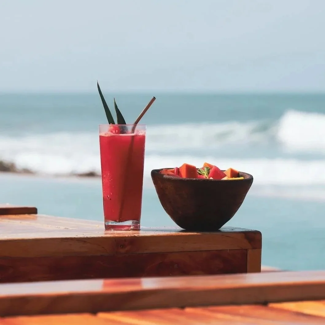 A tall glass of red juice with garnishes, and a wooden bowl of fruit slices on a wooden table with ocean waves in the background.