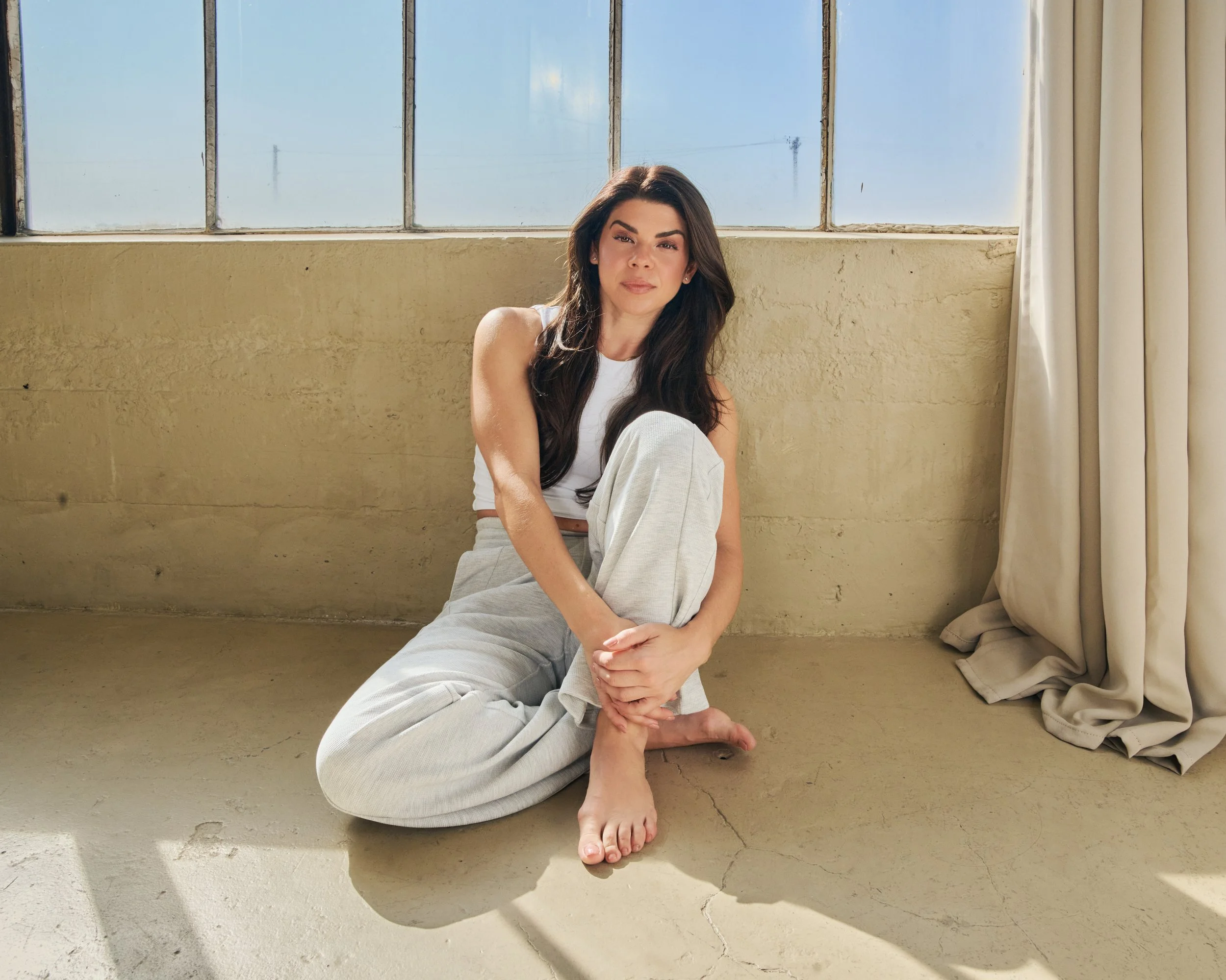 A woman with long dark hair sitting on the floor near a large window, wearing a white tank top and light gray sweatpants, with a beige curtain on the right side, and sunlight casting shadows on the floor.