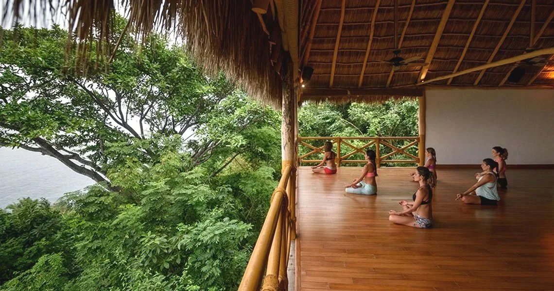 A group of women practicing yoga on a wooden floor terrace surrounded by lush green trees, with a thatched roof overhead.