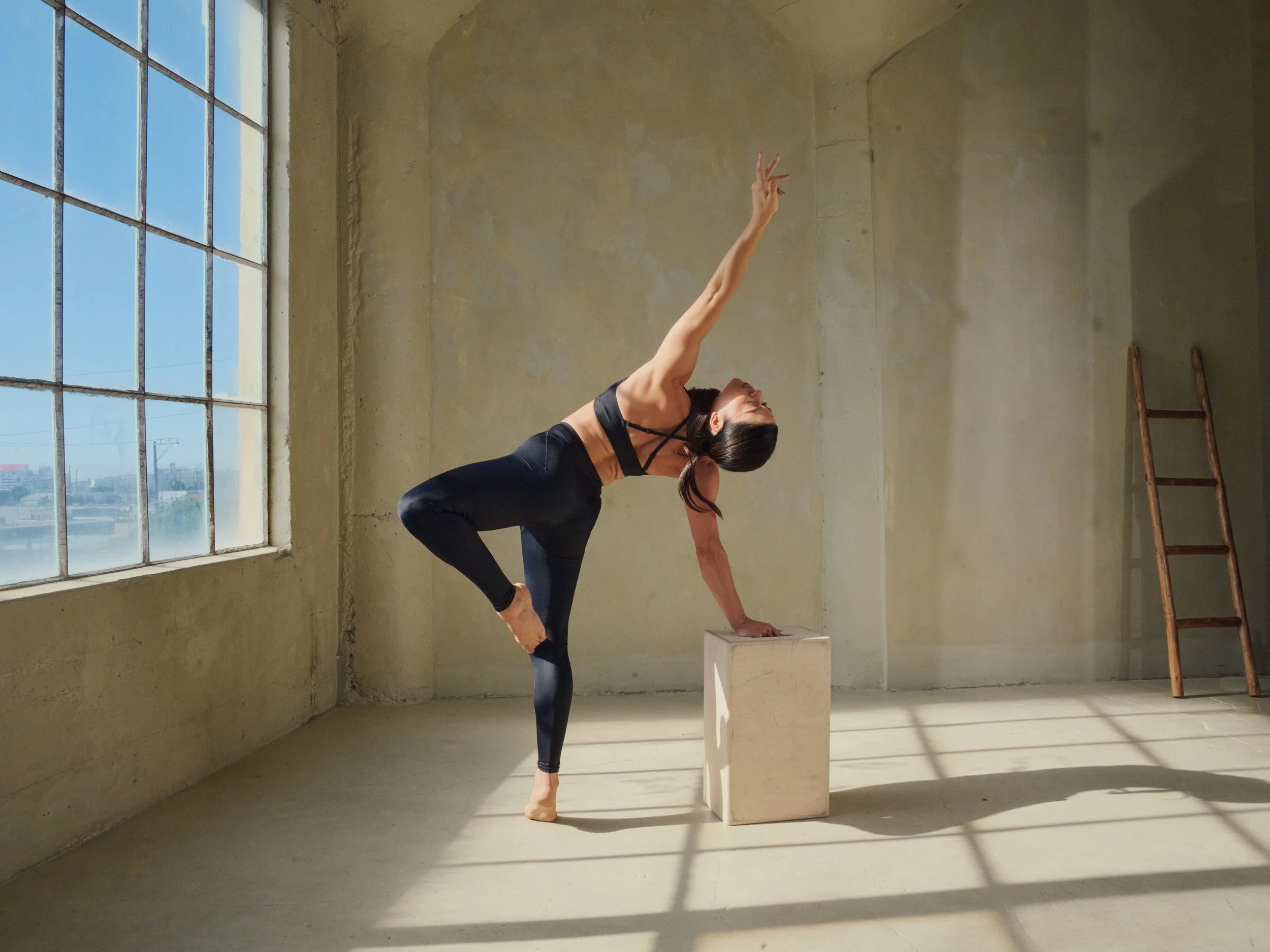 A woman practicing yoga indoors, balancing on one foot with one hand on a block and the other extended upward, in a spacious room with large window and natural light.