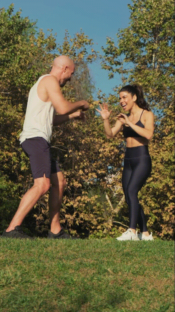 A man and woman outdoors on a grassy area, smiling and playfully interacting with each other, with trees and a clear blue sky in the background.