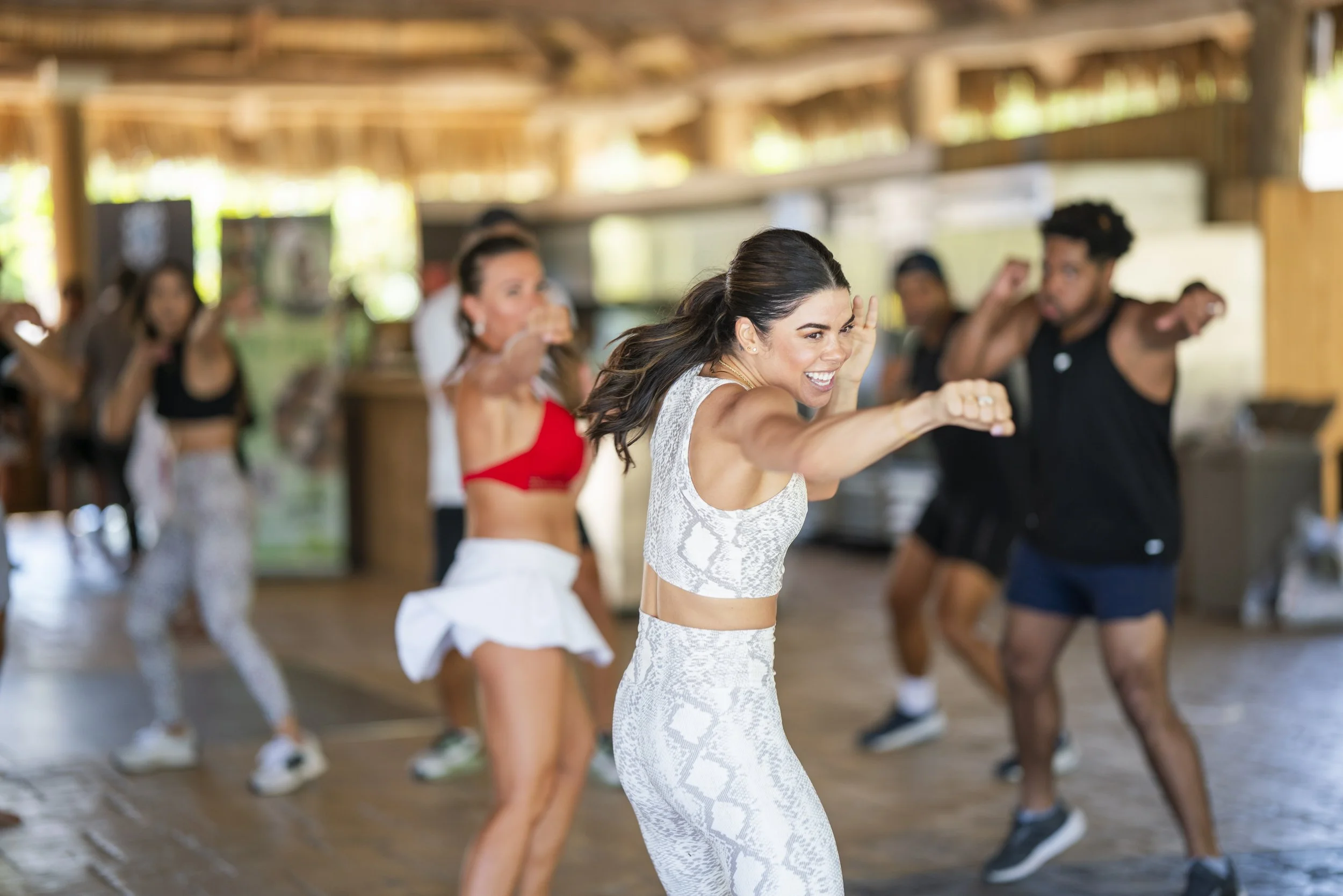 Group of people participating in a dance or aerobic class in a rustic indoor setting, led by a woman in a white patterned workout outfit smiling and reaching forward.