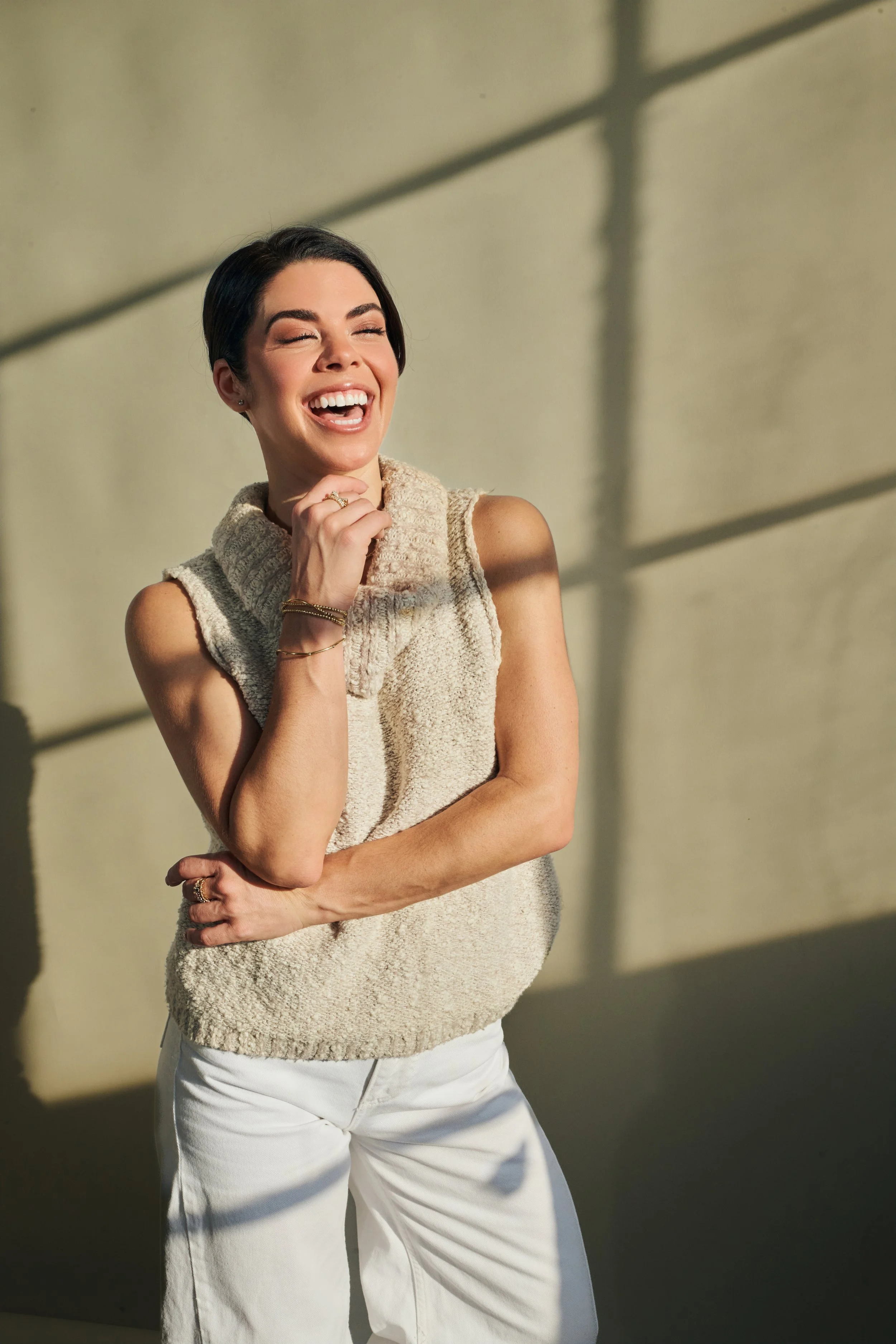 A woman with short dark hair laughing and posing against a wall with sunlight and shadows.