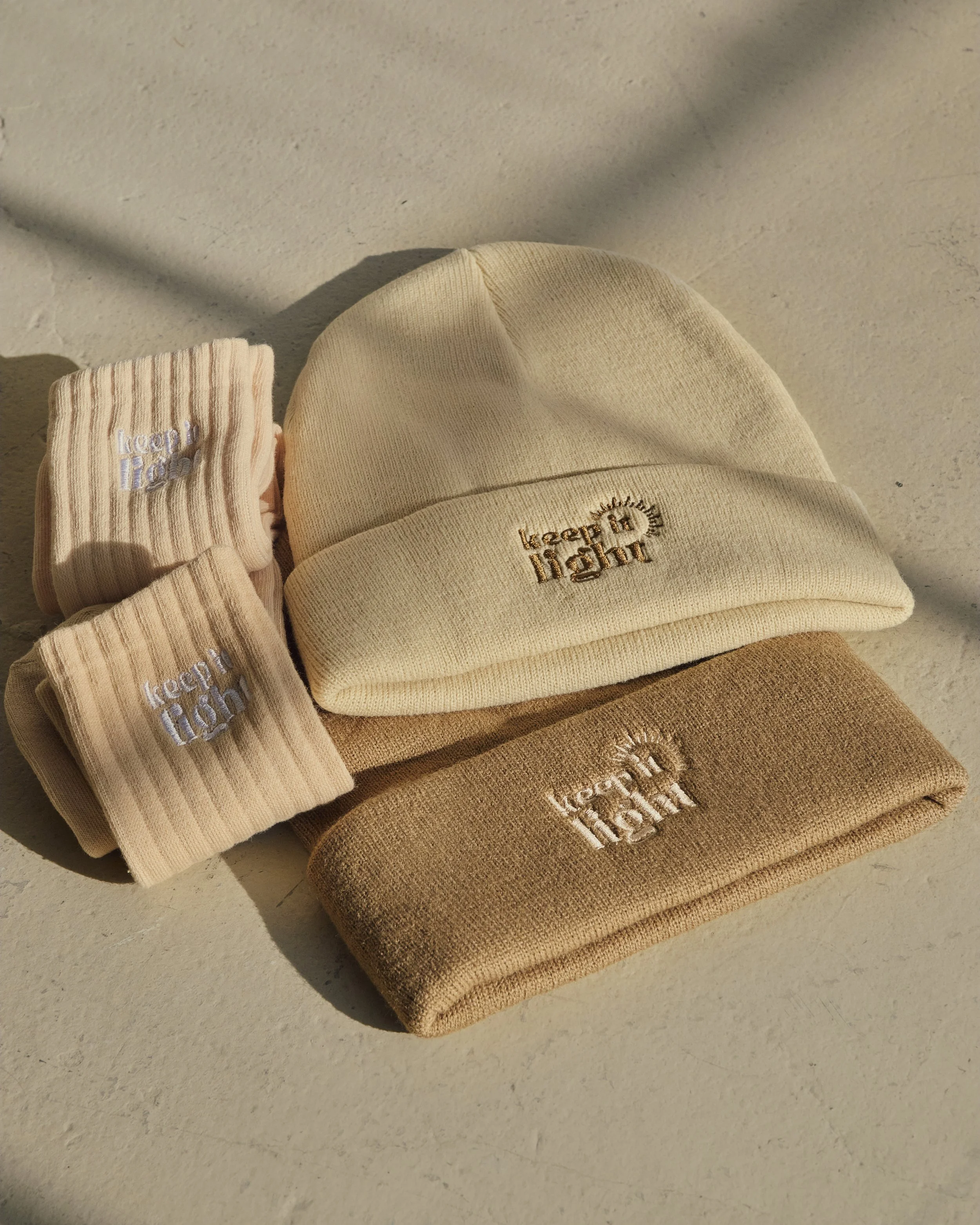 Set of beige and cream-colored beanie hats and socks with embroidered 'Keep it Light' text.