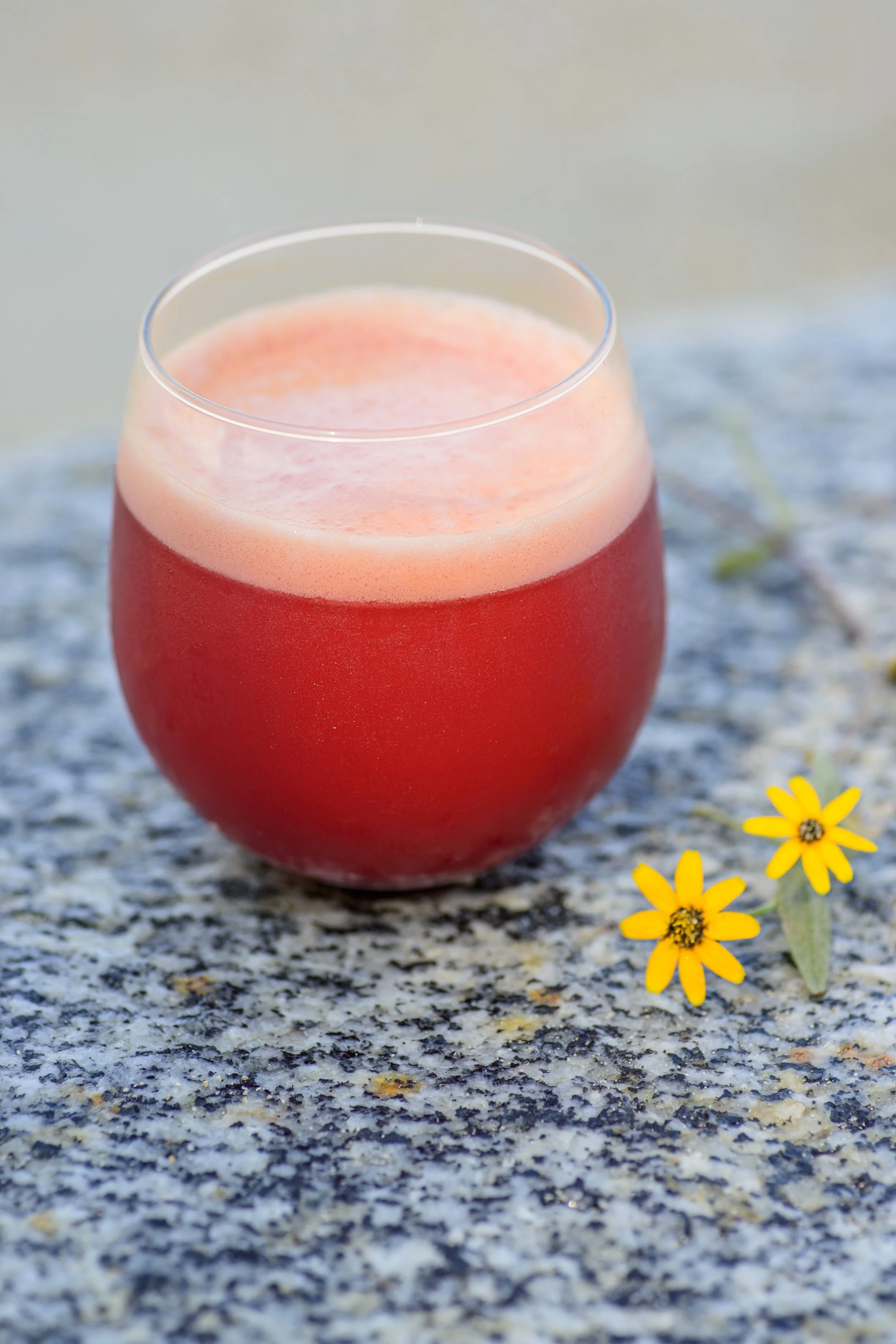 A glass of red, frothy beverage on a granite surface, with two yellow flowers nearby.