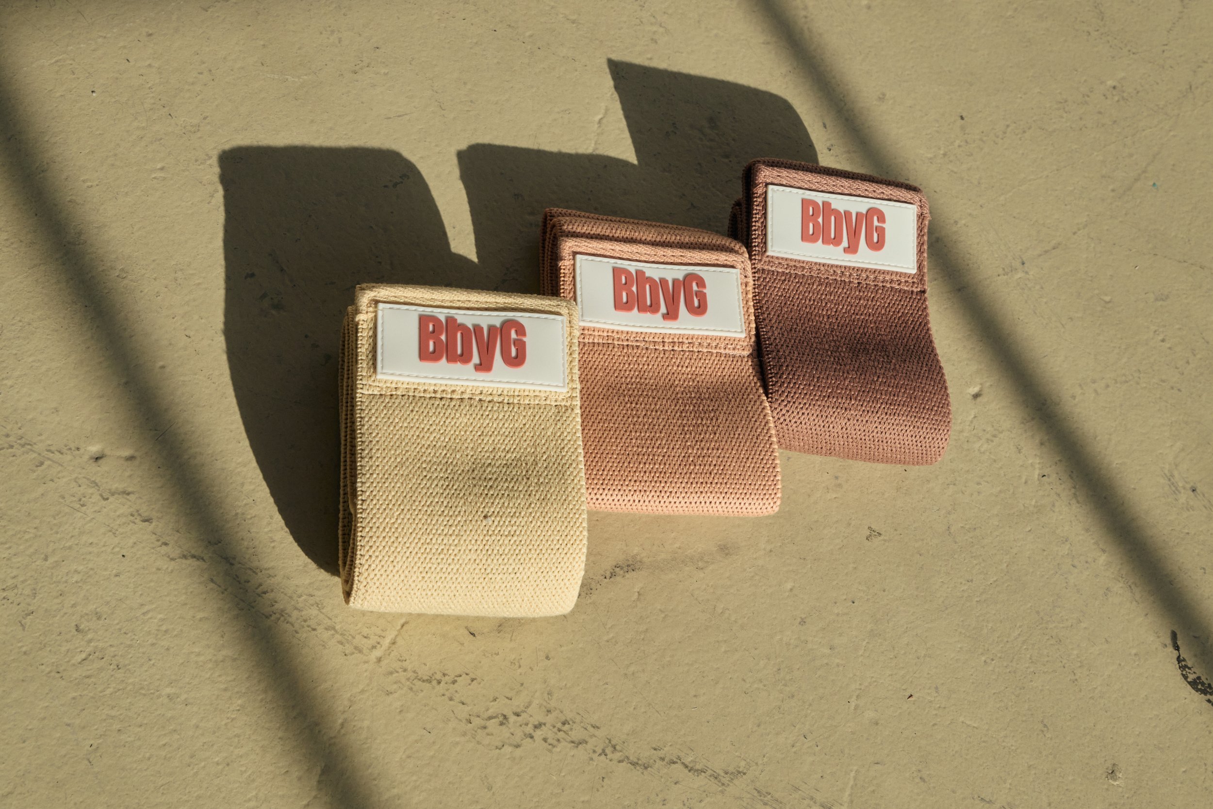 Three pastel-colored elastic hairbands with white labels reading 'BbyG' are lined up on a concrete surface with shadows cast across the background.