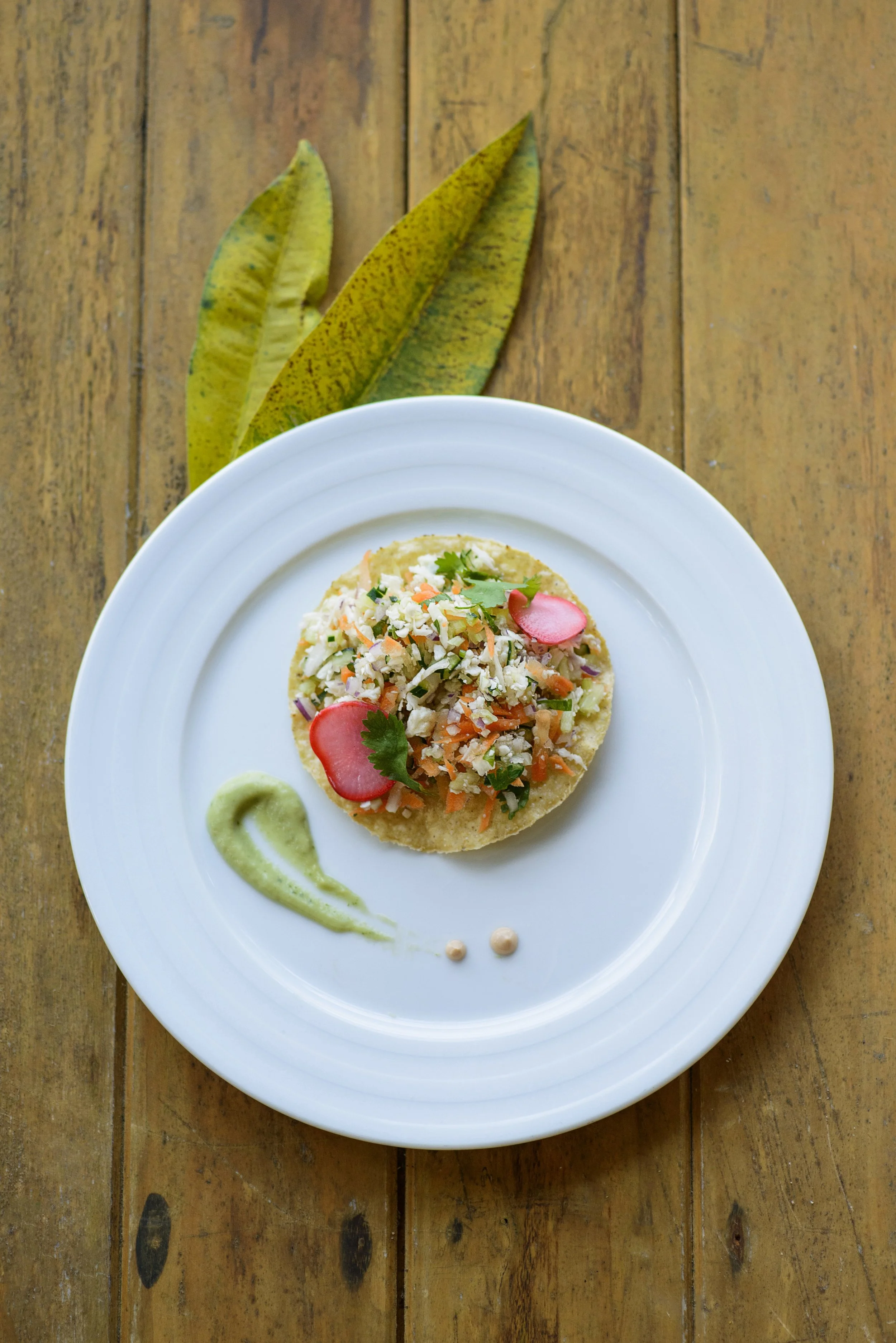 A plate of Mexican street corn topped with chopped vegetables and herbs, garnished with radish slices and cilantro, on a wooden surface with green leaves in the background.
