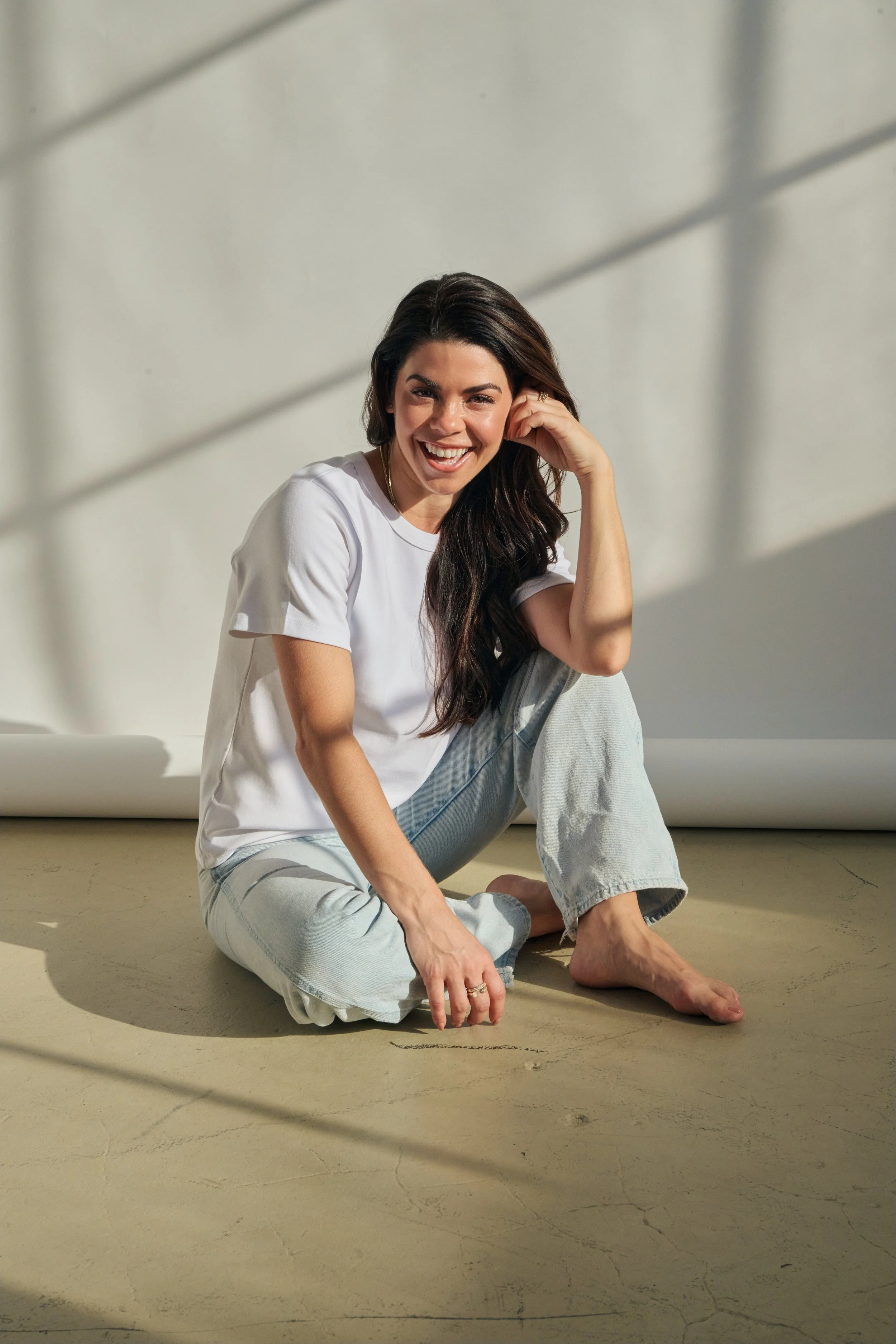 A smiling woman with long dark hair, sitting on the floor in casual white t-shirt and jeans, with sunlight and shadows on a plain background.