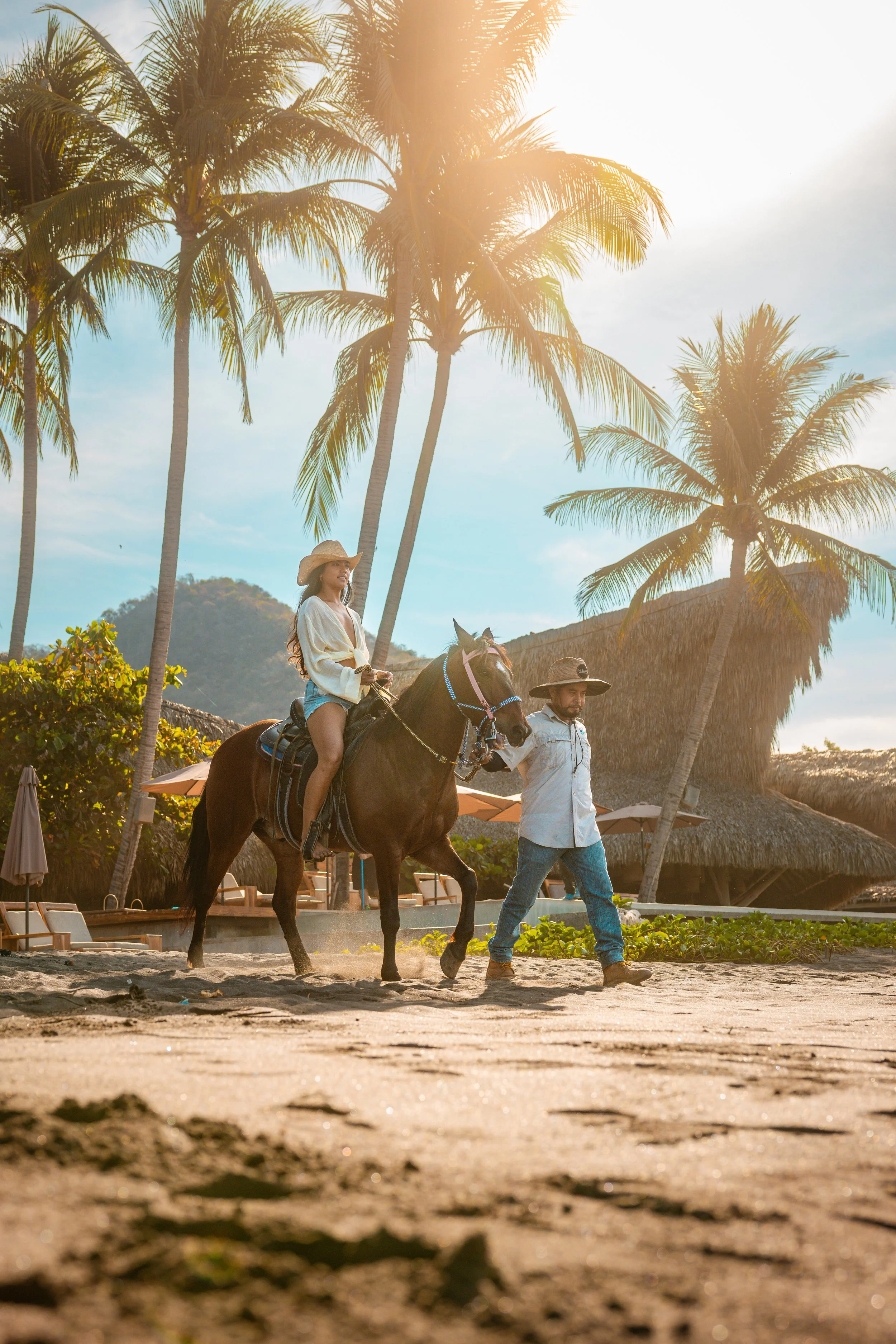 A woman riding a horse on a beach with palm trees, a building with thatched roof, and a man walking beside her, during sunset.