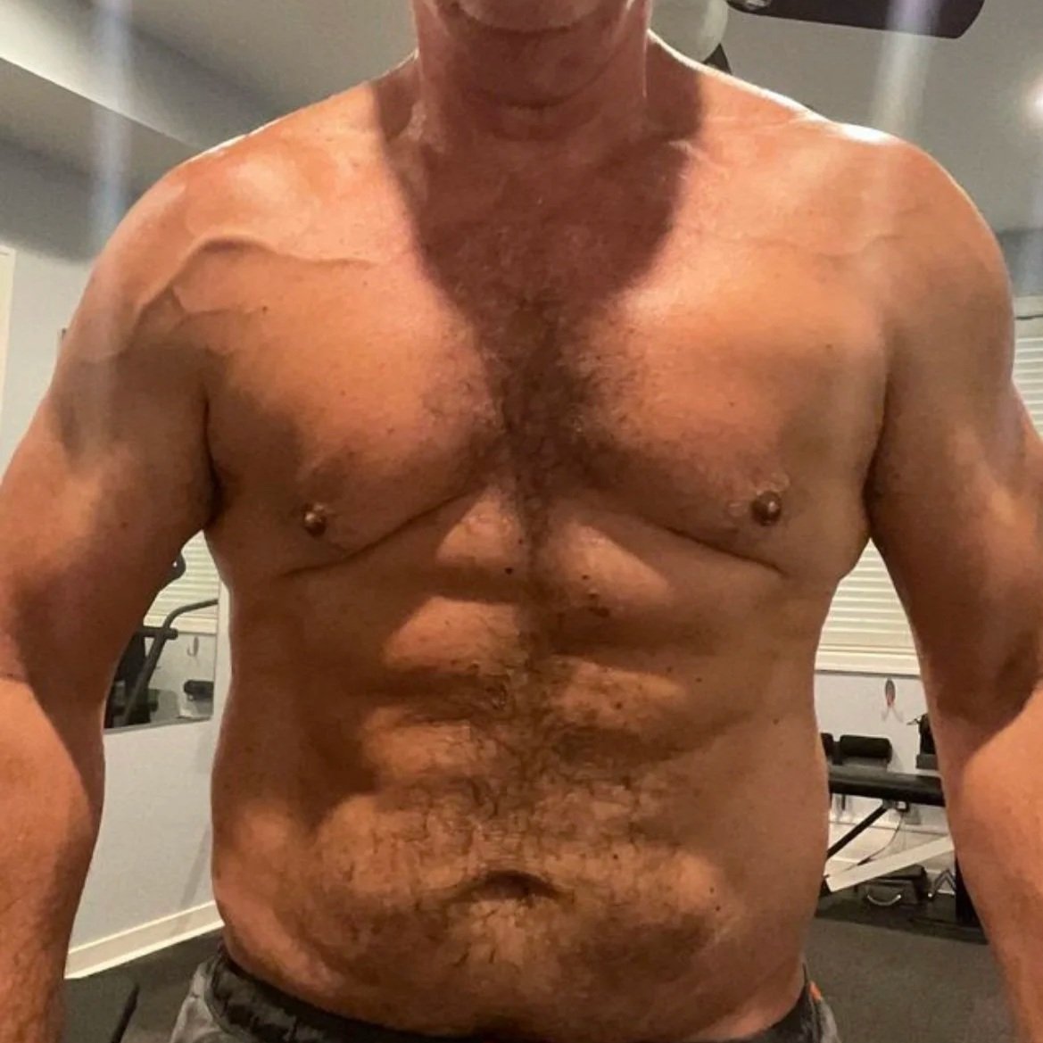 A shirtless man with a muscular build standing in a home gym, with various exercise equipment visible in the background.