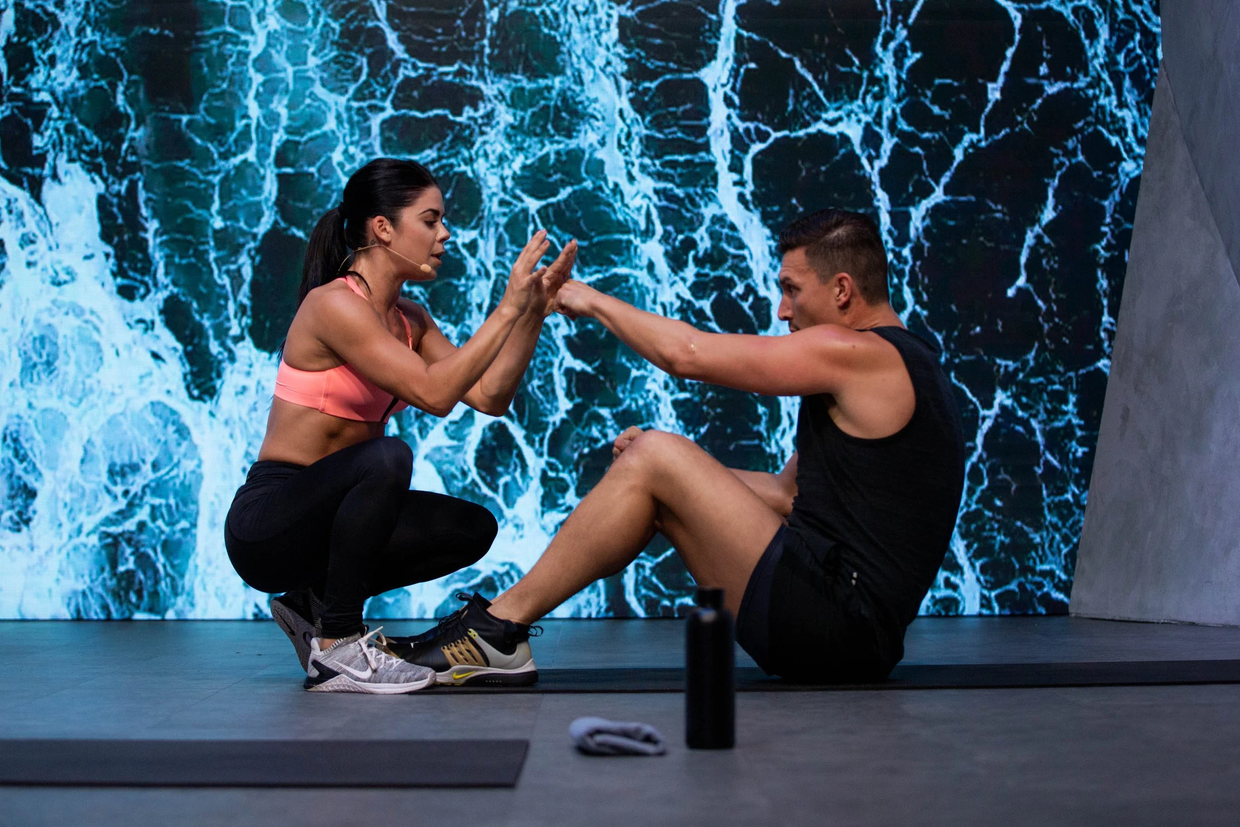 A woman in athletic gear is assisting a man with an exercise on a workout mat, with a digital water background behind them.