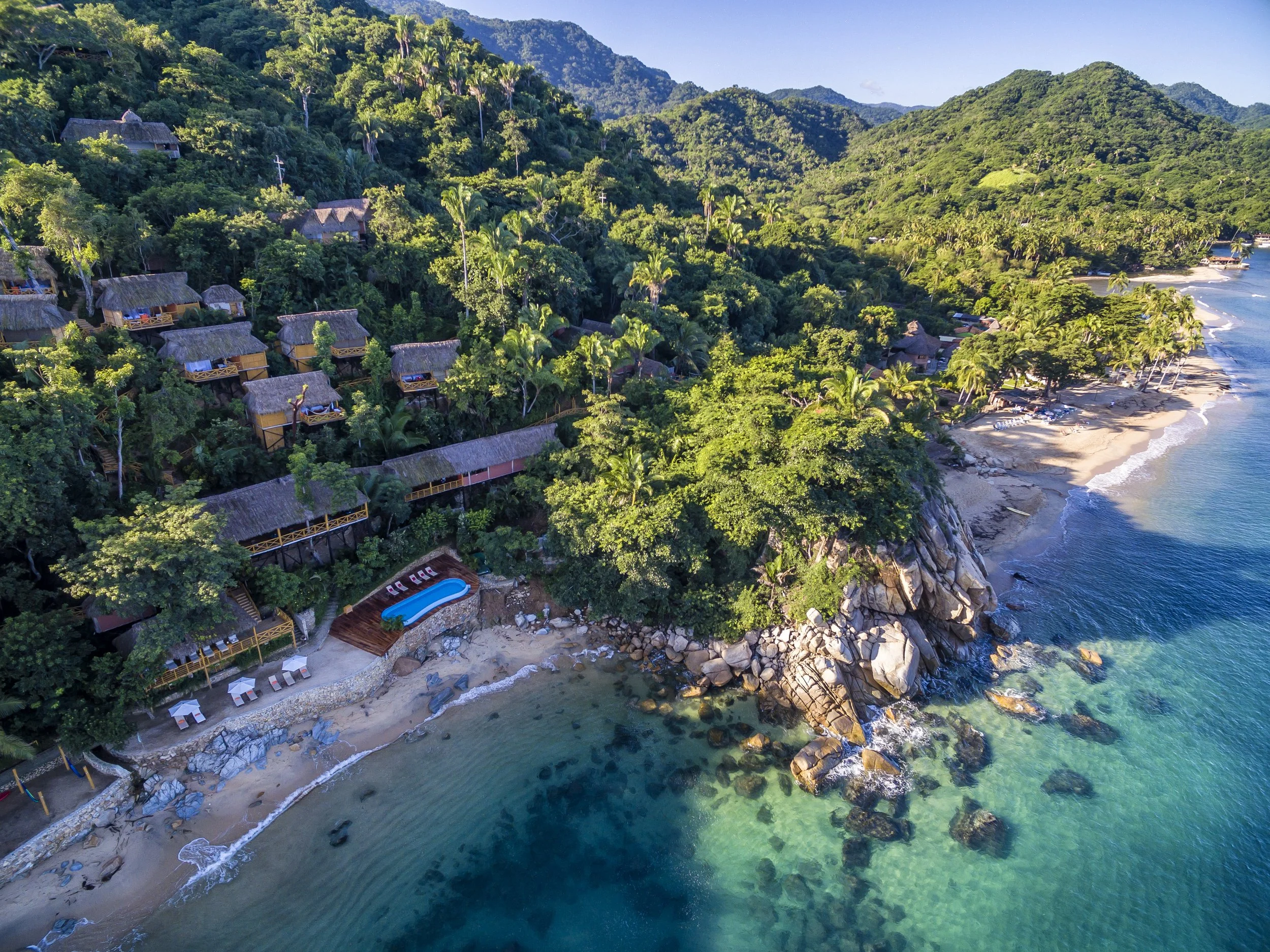 Aerial view of a tropical resort on a hillside, featuring multiple treehouse accommodations with thatched roofs, a swimming pool, a sandy beach, and clear turquoise water.