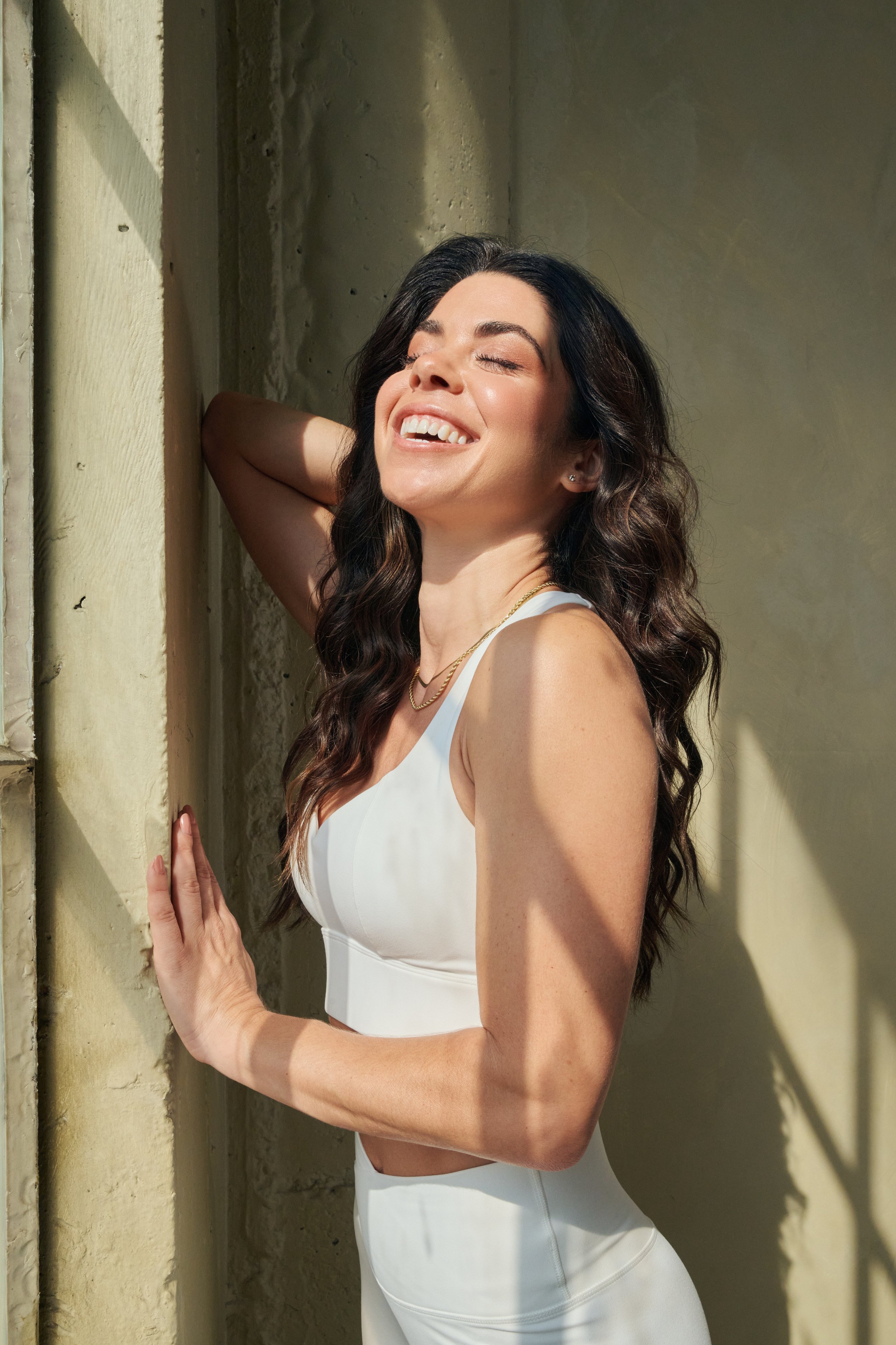 A woman with long dark hair, smiling with eyes closed, leaning against a textured wall in sunlight, wearing a white sleeveless top and gold jewelry.