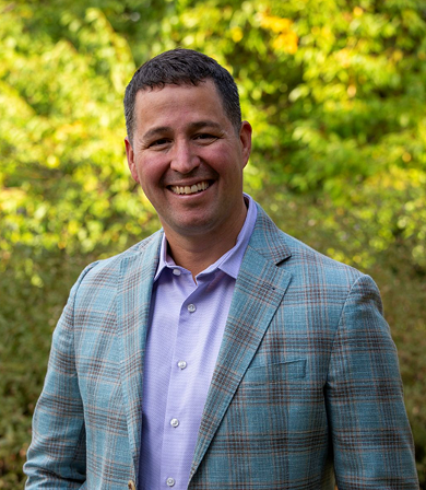 A picture of Andrew Martinez-Alegria, founder of Sound Talent Strategies, in a purple shirt with a blue plaid sport coat in a natural background setting.