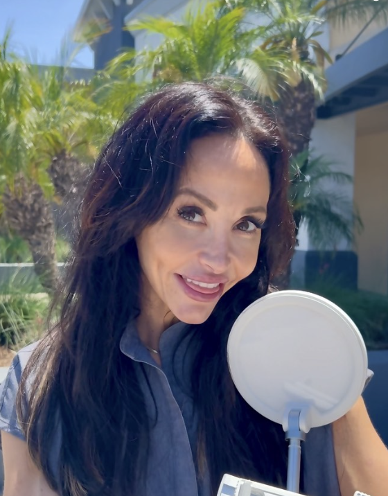 A woman with dark brown hair wearing a gray top, smiling and standing outdoors with palm trees in the background.