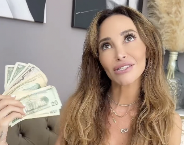 A woman with long, wavy hair holding a fan of 20-dollar bills, looking up with a thoughtful expression in a modern office.