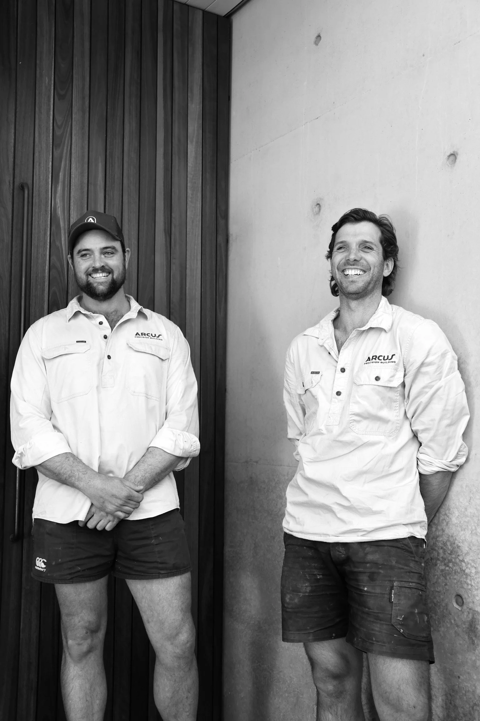 Co-owners Toby Williams and Harry Capel, smiling in Arcus Projects work uniform standing against a wooden and concrete wall.