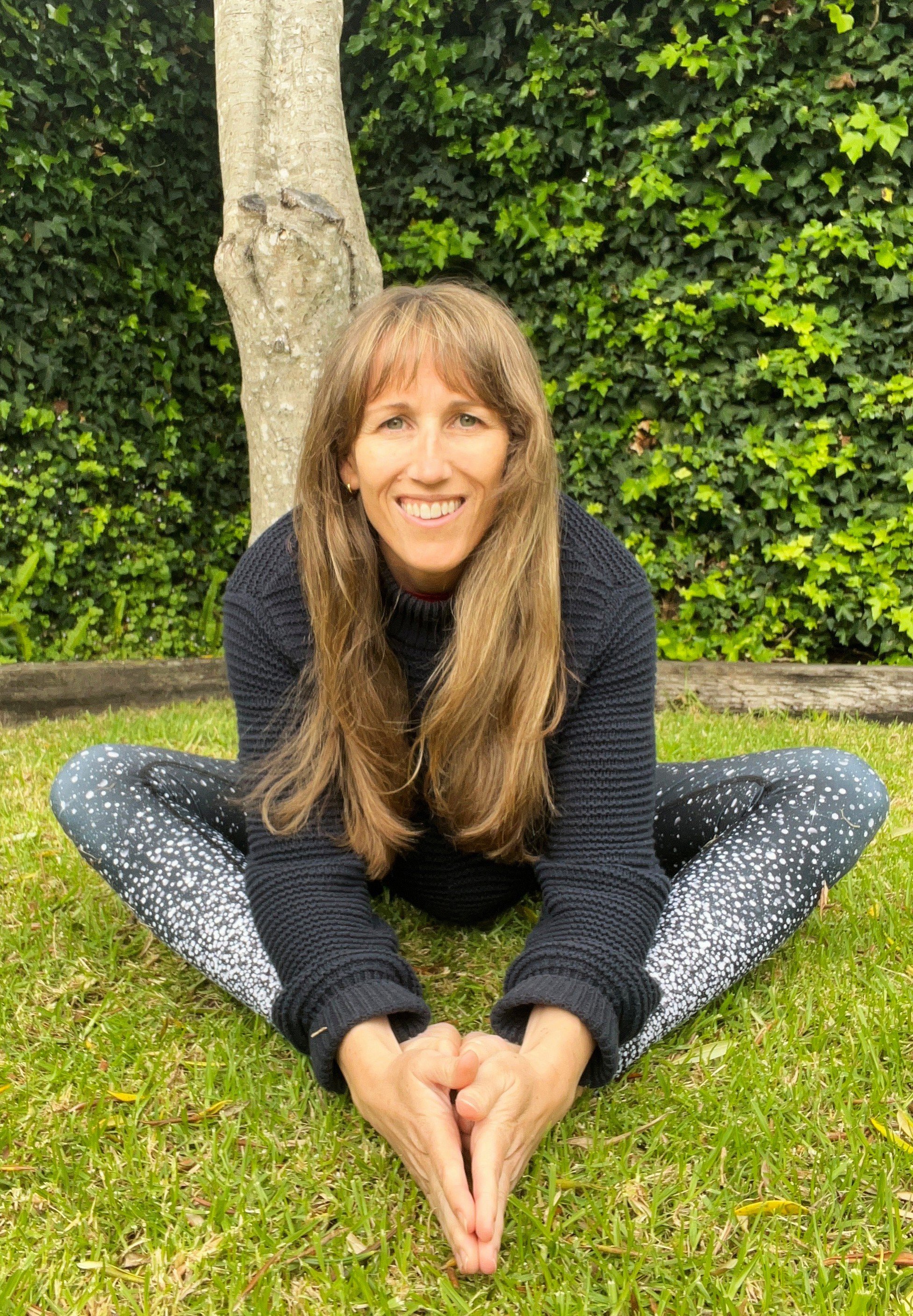 A woman with long brown hair sitting cross-legged on the grass in front of a tree and green shrubbery, smiling at the camera.