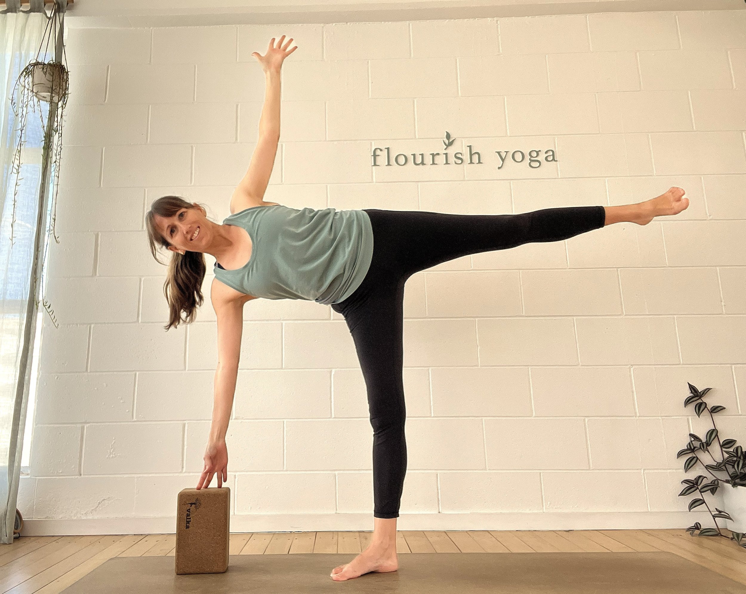 A woman practicing yoga indoors, balancing on her right leg with her left leg extended horizontally, one arm reaching down toward a yoga block, and the other arm extended upward. The wall behind her has the words "flourish yoga" with a small leaf icon, and there is a potted plant to her right.