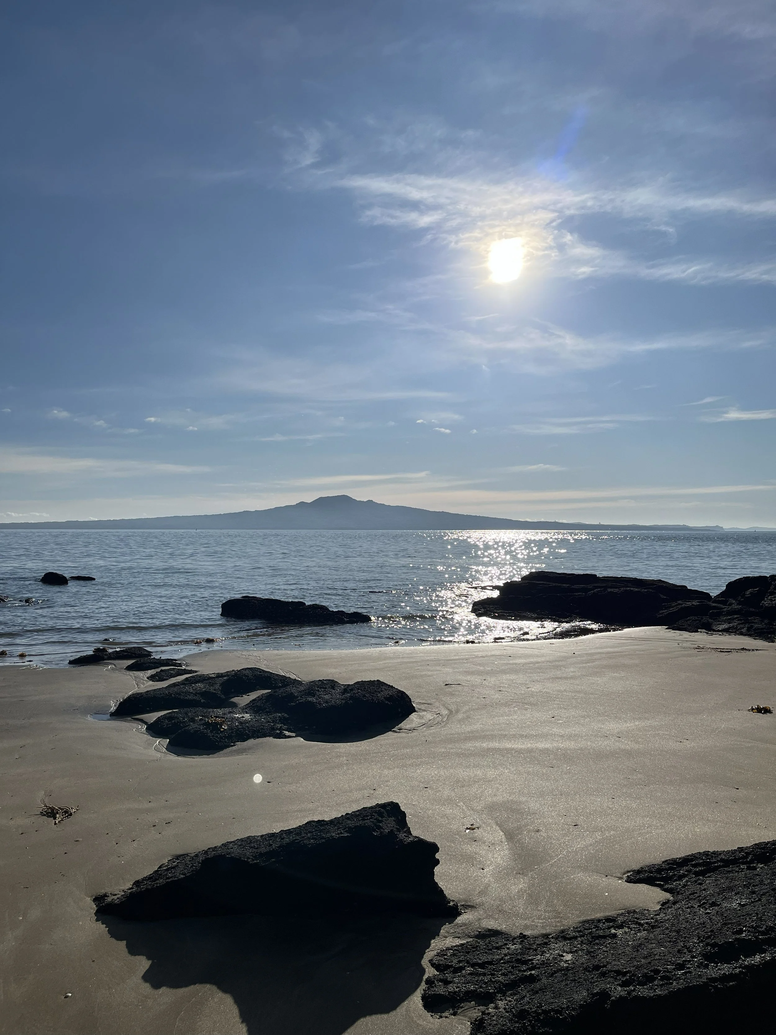A scenic beach with rocks on the sandy shore, calm water reflecting sunlight, distant landmass or island, and a clear sky with the sun shining.