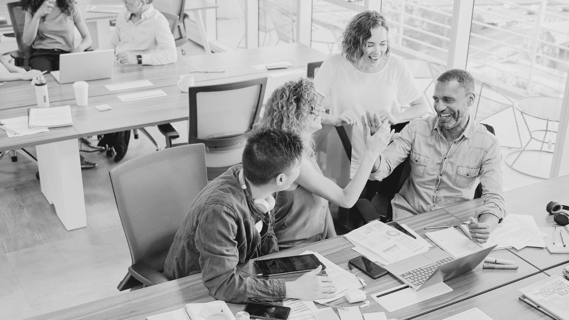A group of five people in a modern office, two are sitting at a table working on laptops and papers, while three are standing nearby, one woman appears to be congratulating a man. Everyone is smiling and engaging in a positive interaction.