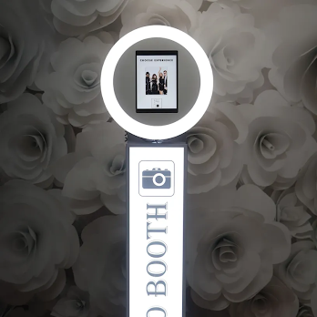 Photo of a photo booth sign with a digital screen displaying a group photo and a camera icon, set against a background of large white roses.
