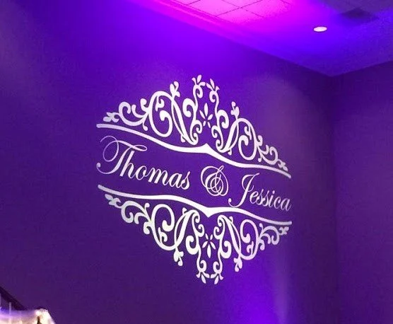Wall decoration with the names Thomas and Jessica in elegant script, surrounded by ornate floral designs, lit with purple lighting.