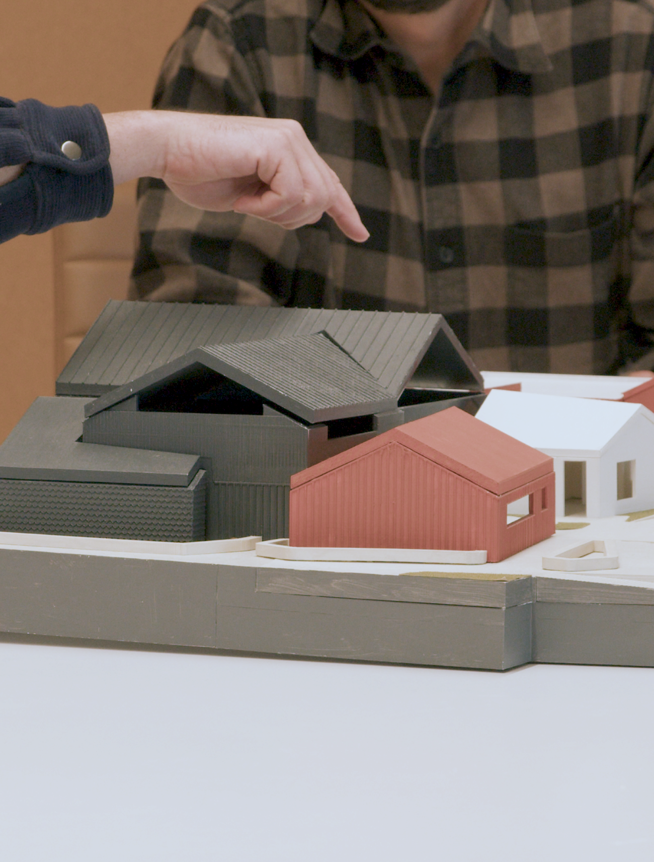 A person pointing at a model of various house buildings, including black, red, and white structures, on a table.