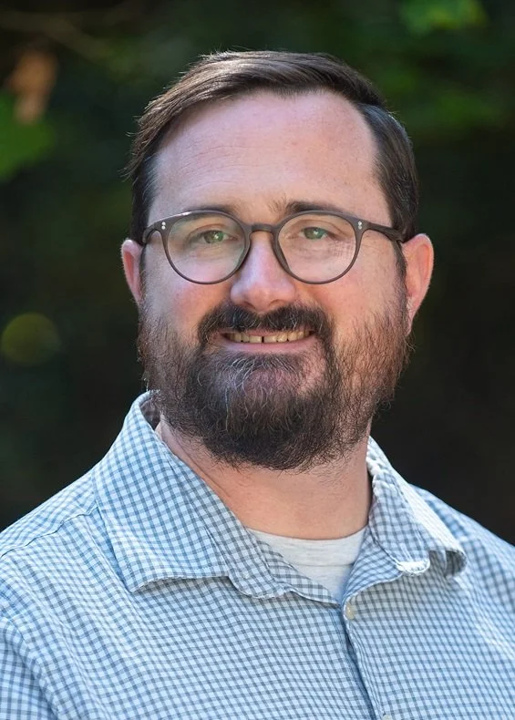A man with glasses and a beard, smiling, wearing a checkered shirt outdoors.