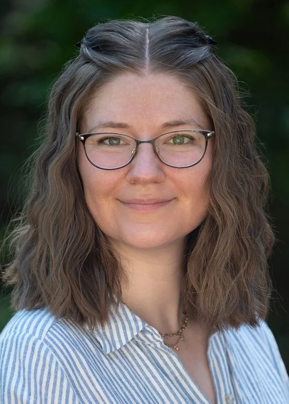 A woman with shoulder-length wavy brown hair, glasses, and a gentle smile, standing outdoors with blurred greenery in the background wearing a blue and white striped shirt.