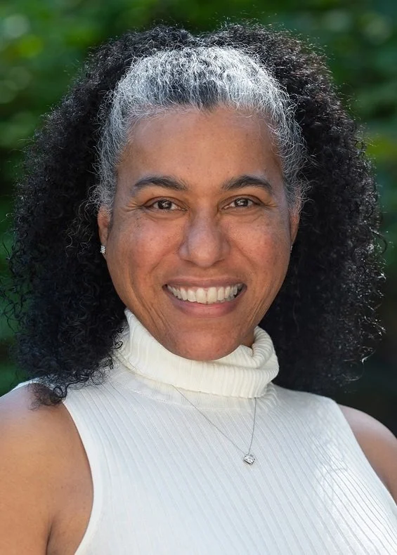A woman with curly black and gray hair smiling outdoors, wearing a white sleeveless turtleneck and a necklace.
