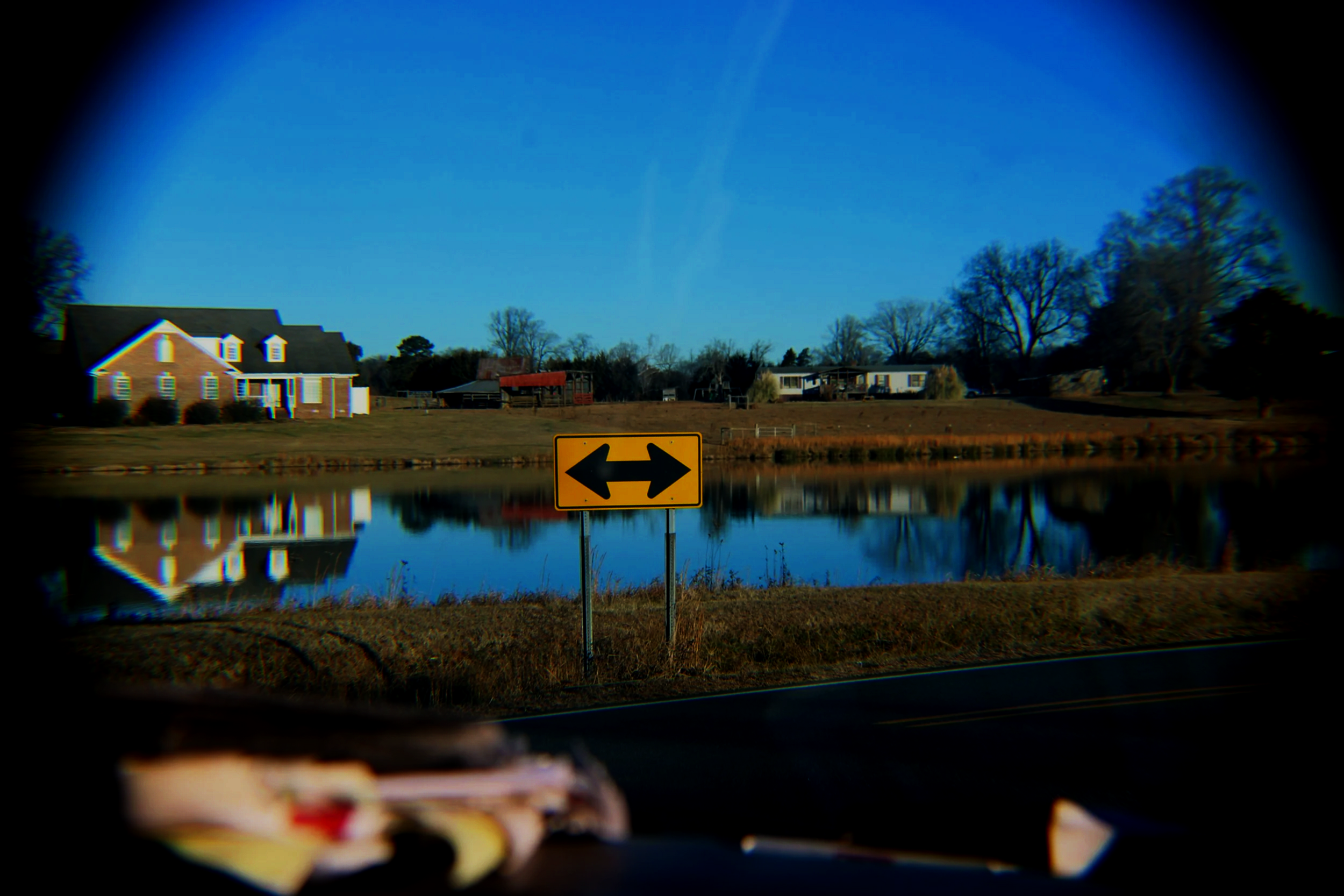 A view through a car's side mirror showing a yellow traffic sign with black arrows pointing left and right, reflecting houses and trees across a body of water under a clear blue sky.