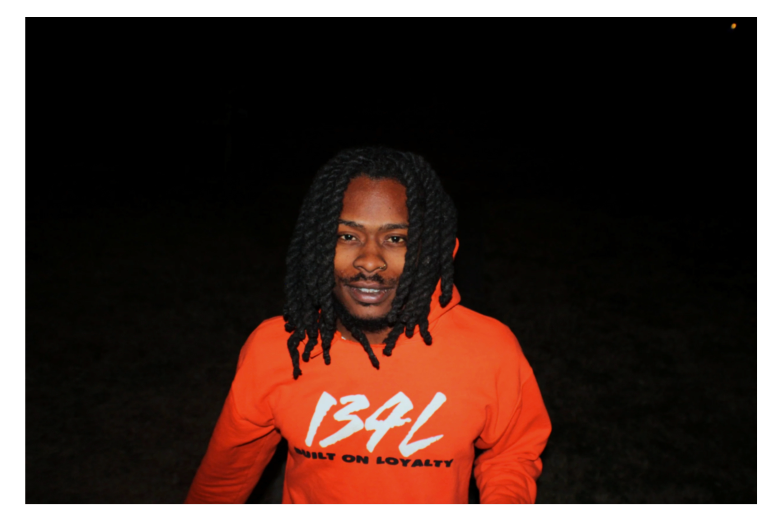 A man with black, textured dreadlocks smiling at the camera. He is wearing an orange hoodie with white and black text that reads '134L Built on Loyalty.' The background is black.
