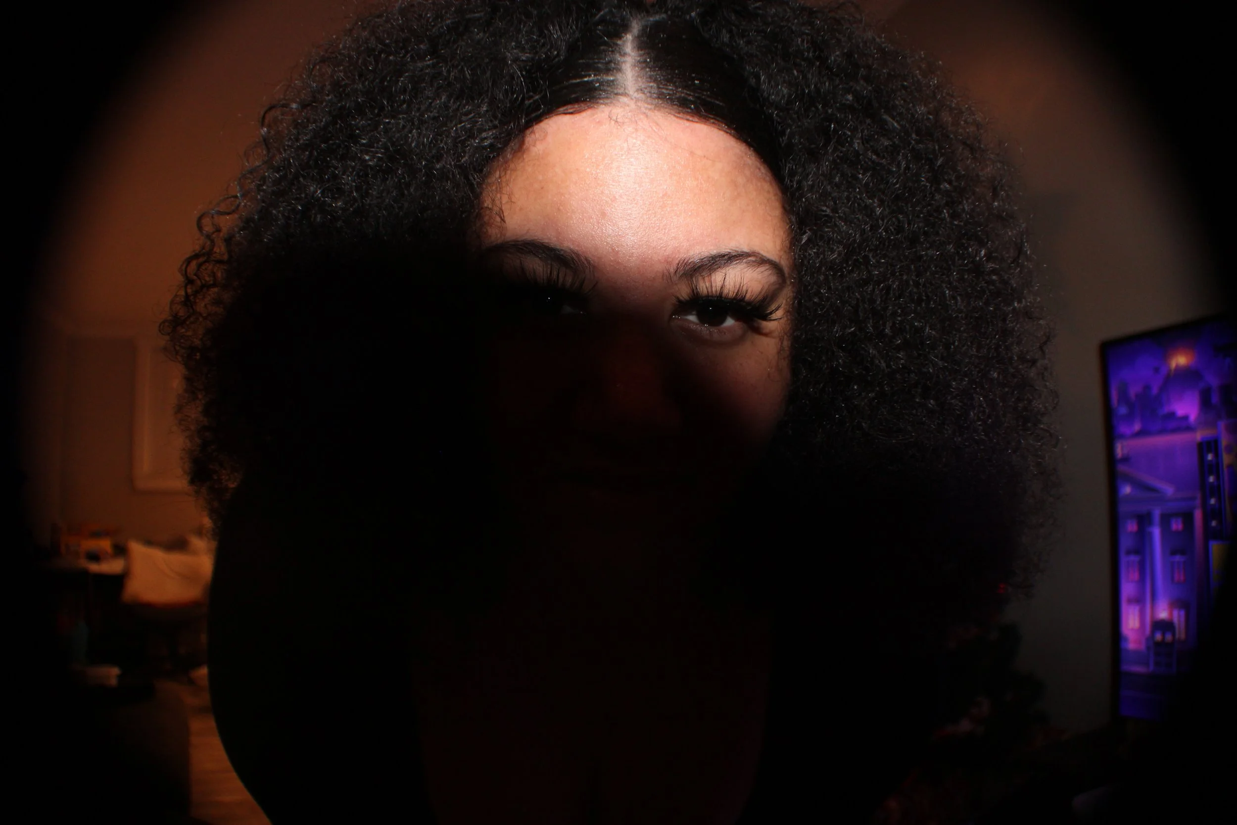 A woman with curly hair is illuminated by a light source in a dark room, with a TV screen showing colorful images in the background.