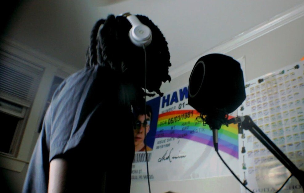 A person with dreadlocks wearing headphones and a black shirt speaking into a microphone in a room with a computer monitor and a colorful sign in the background.