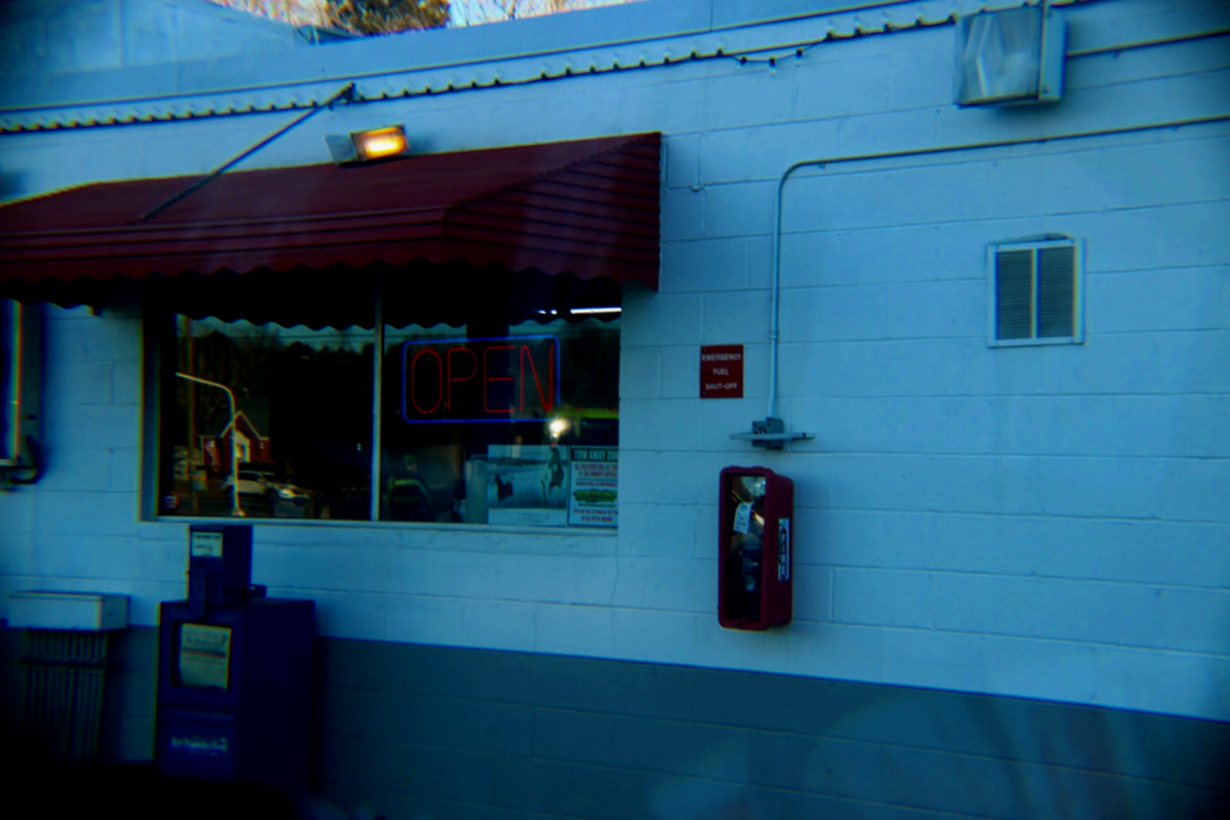 The exterior of a store with a large window displaying a neon 'OPEN' sign. There is a red awning above the window, and a red emergency pull box mounted on the white brick wall to the right of the window. A small window is above the pull box, and a ceiling light illuminates the store entrance.
