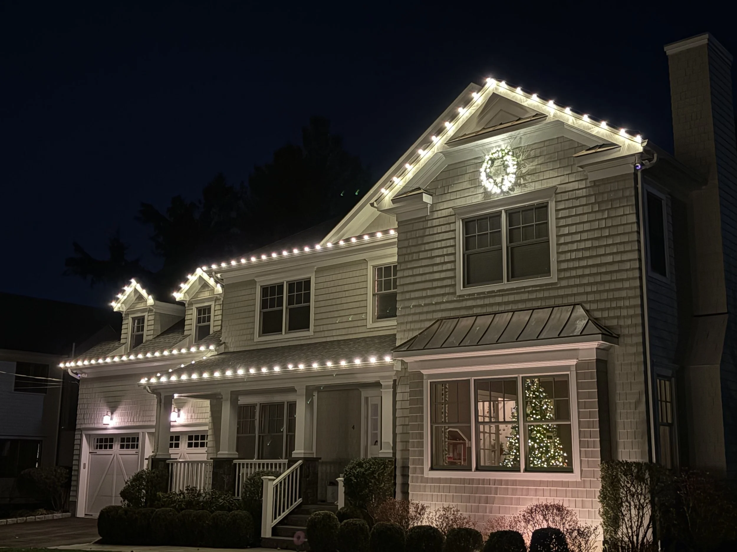 A two-story house with beige siding and white trim decorated for Christmas. The roof is lined with white Christmas lights, and a wreath hangs on the gable. There is a Christmas tree with lights inside the house visible through the front window, and the yard has neatly trimmed bushes and small trees.