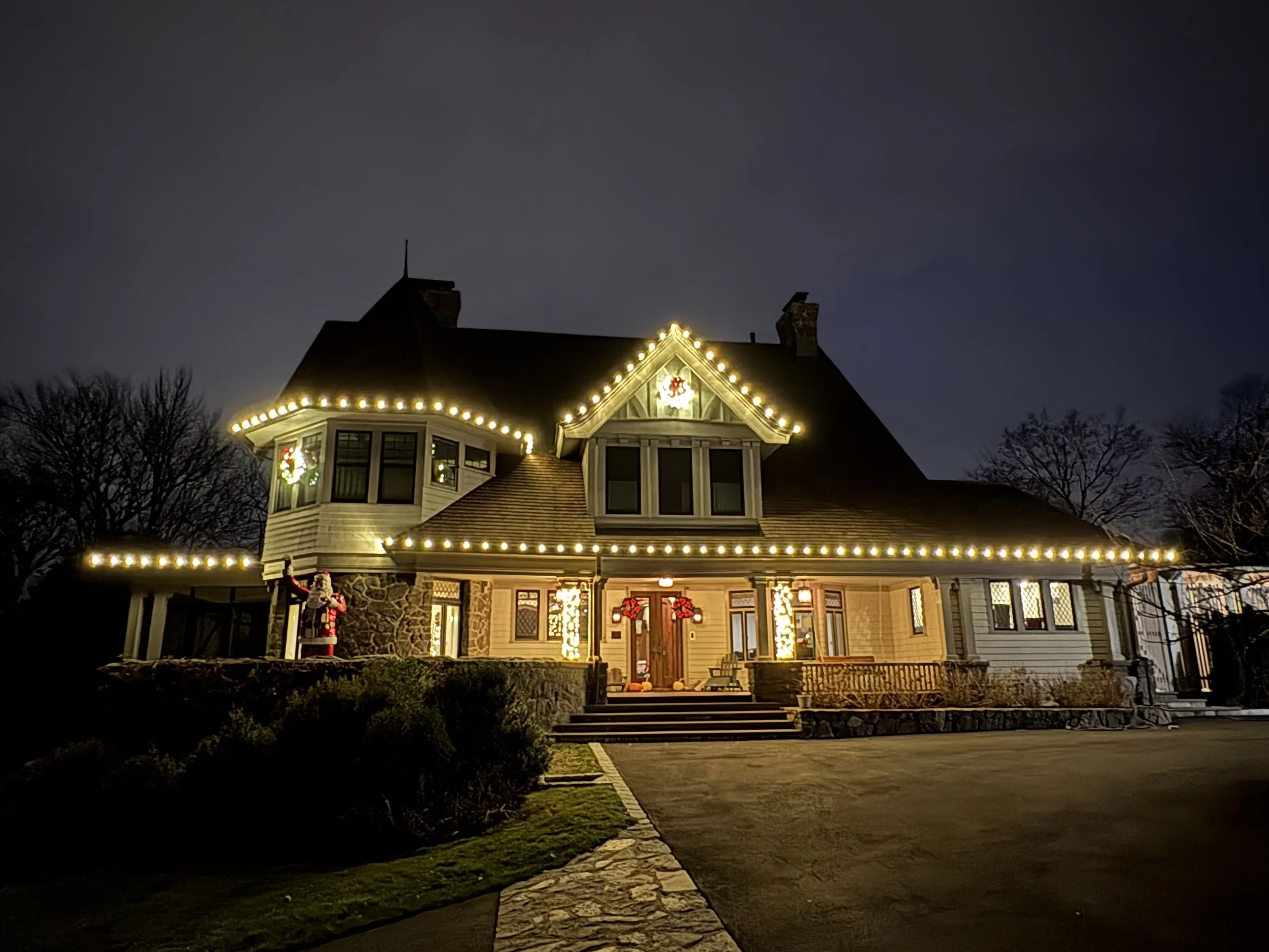 A house decorated with string lights and festive holiday decorations, including wreaths and a Santa figure, under a dark night sky.