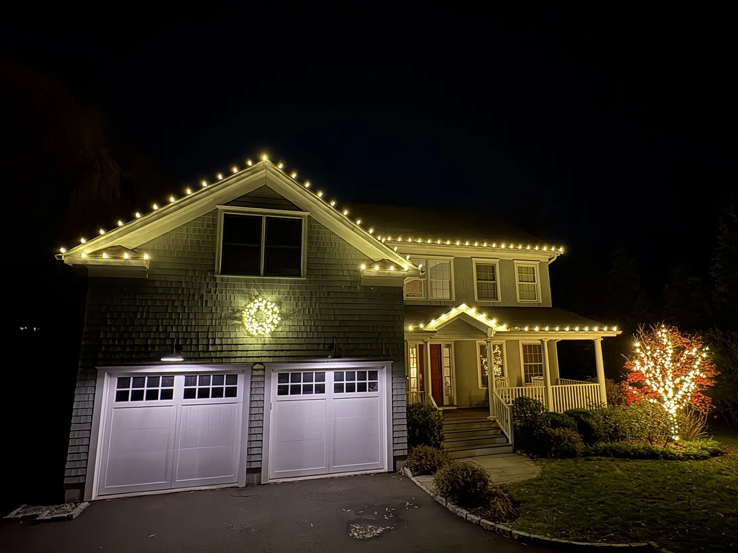 A house decorated with outdoor Christmas lights at night, including a wreath on the wall, and Christmas lights outlining the roof and porch area.