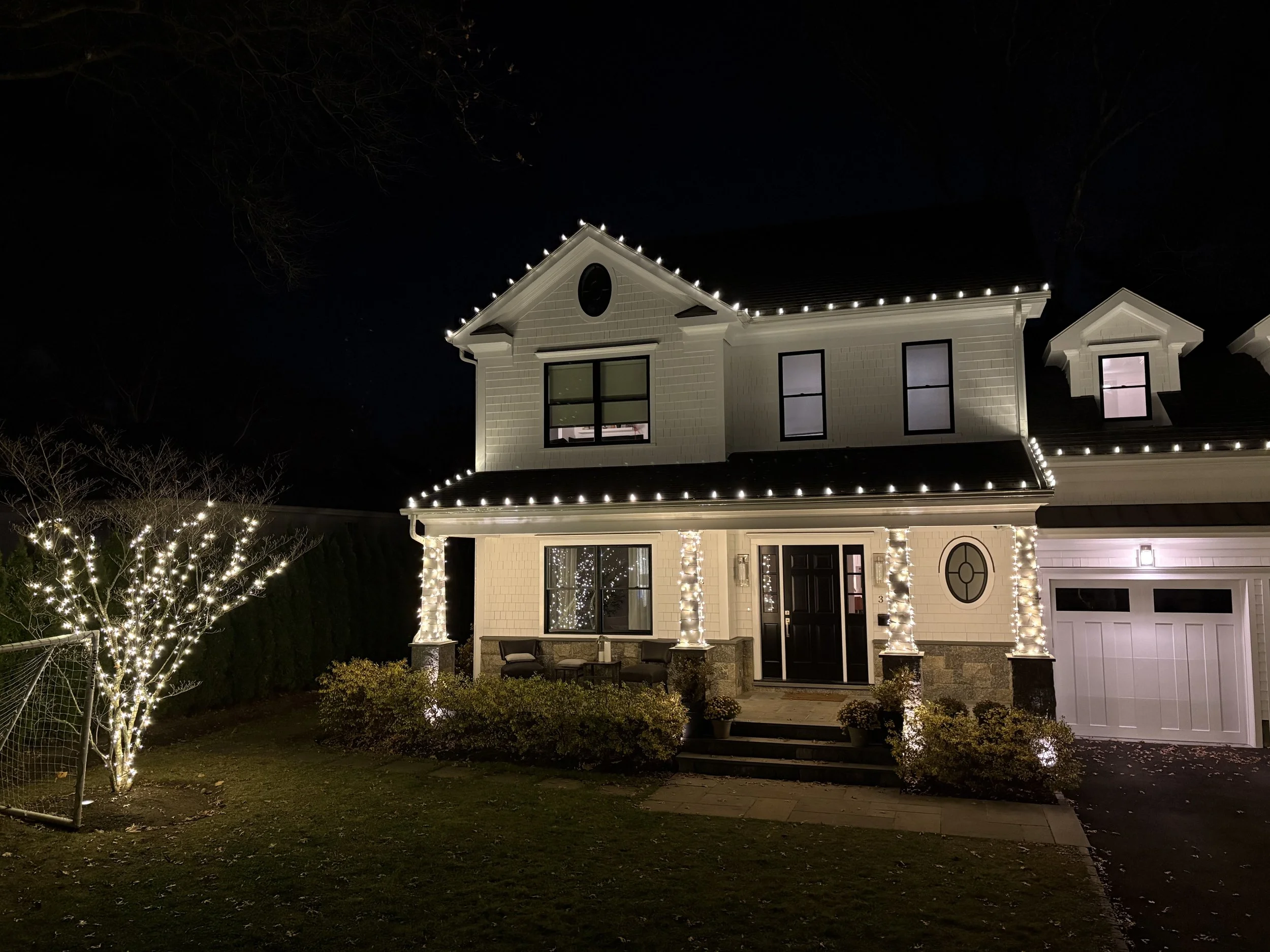A two-story house decorated with white Christmas lights outlining the roof and porch. The house has a black front door, large windows, and a small front porch with chairs. The yard has some bushes and a lit tree on the left side.