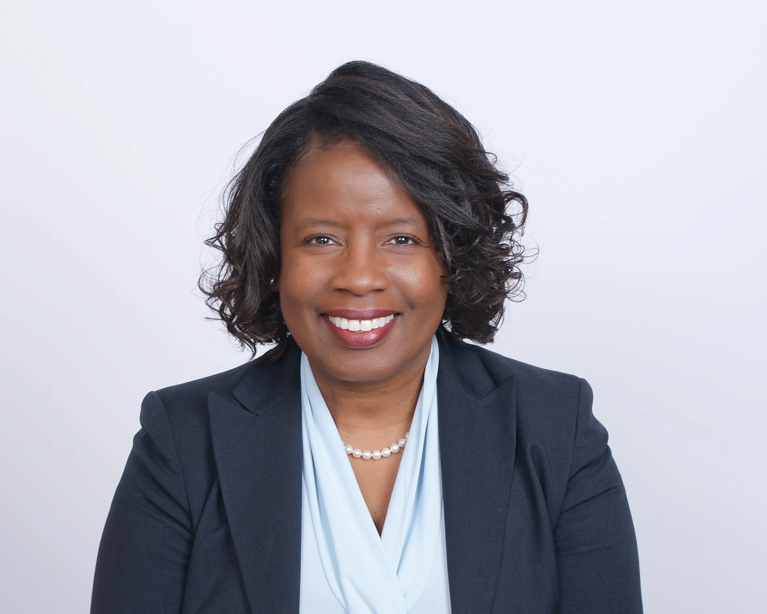 Professional woman smiling, wearing a dark blazer and light blue blouse, with a pearl necklace, against a plain white background.