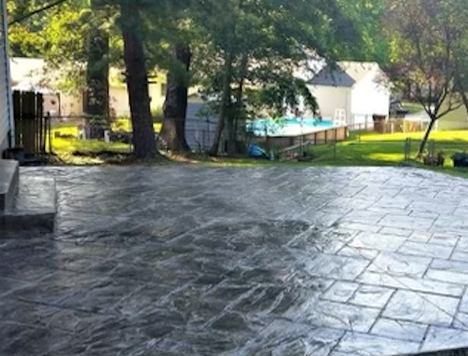 Backyard with stone patio, trees, a swimming pool, and a white shed or small building in the background.