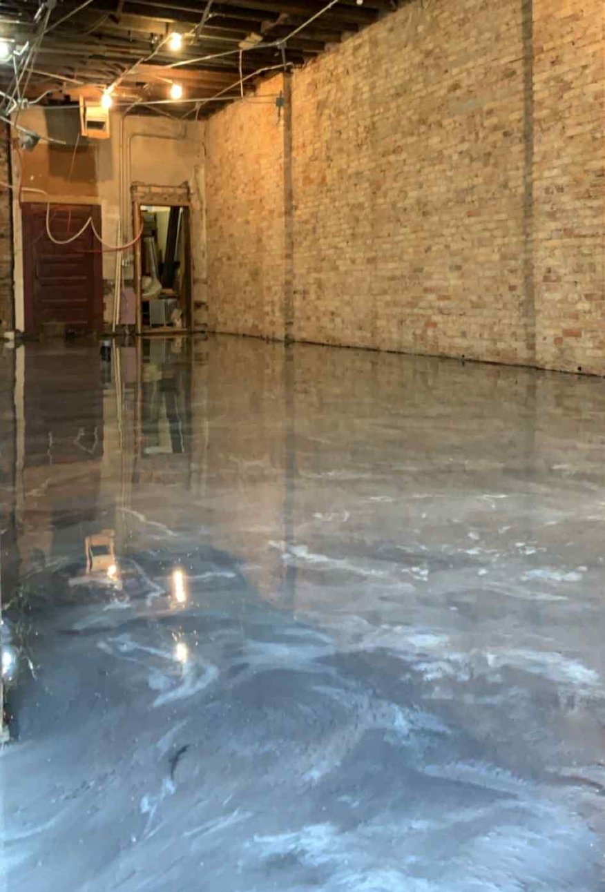 An unfinished interior space with a brick wall, exposed ceiling beams, and a wet concrete floor.