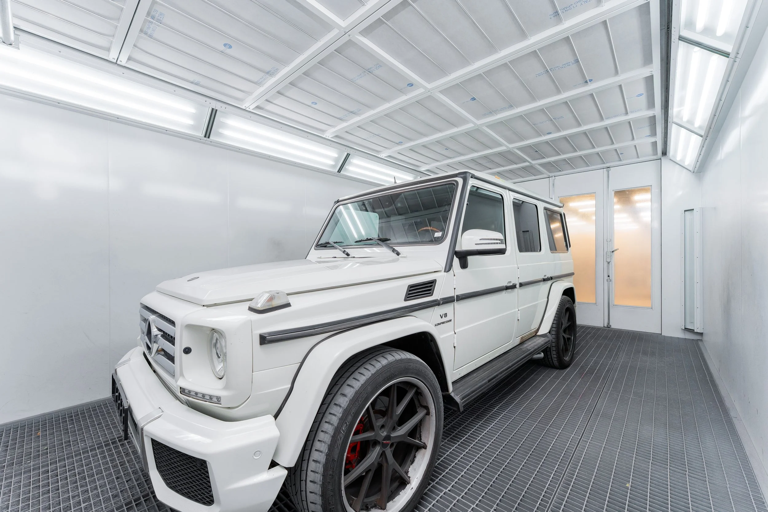 A white off-road vehicle, possibly a Mercedes-Benz G-Class, parked inside a clean, bright paint booth with white walls and ceiling, fluorescent lighting, and a grated metal floor.