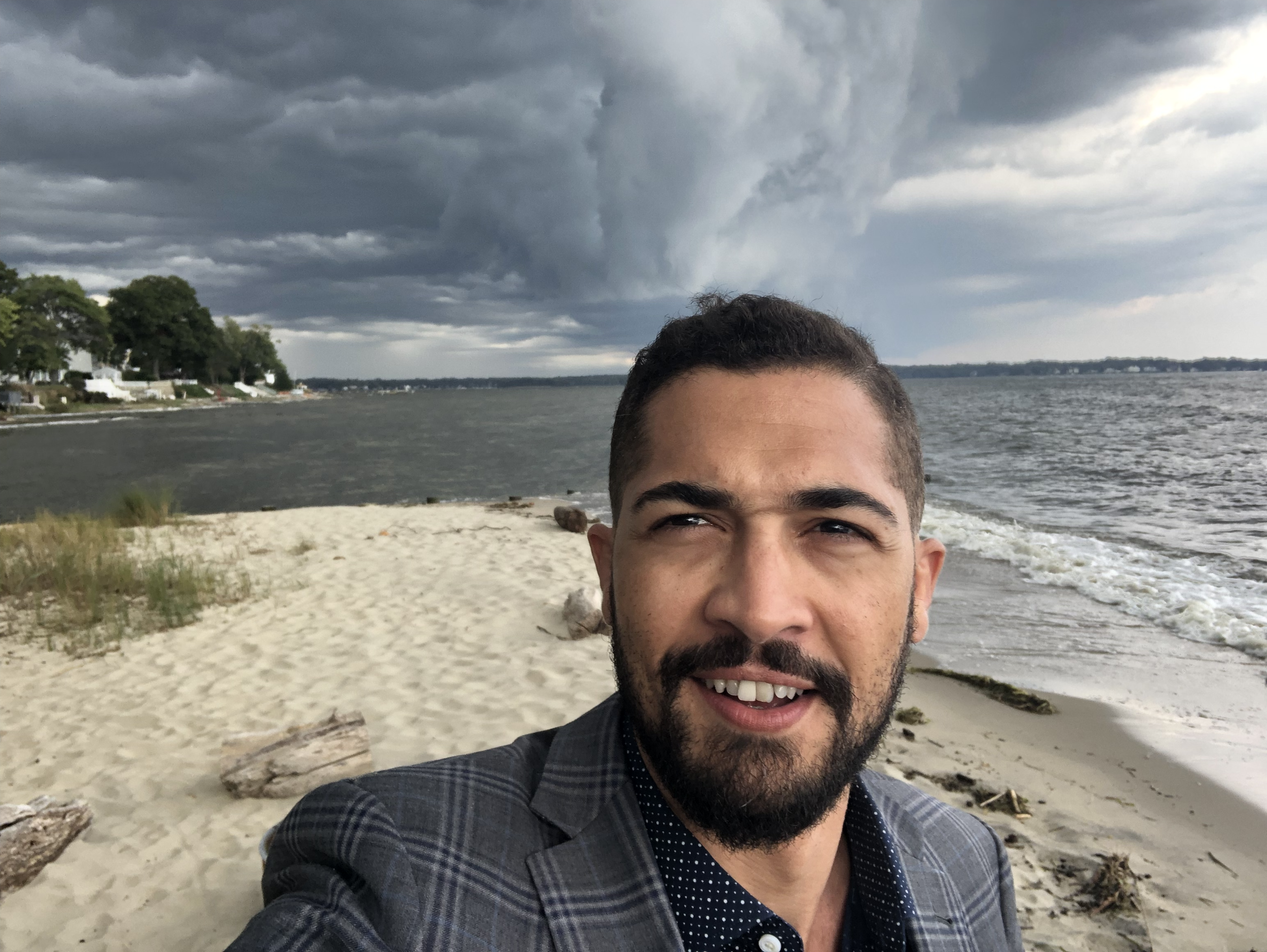 A man taking a selfie on a beach with cloudy sky and a storm approaching over the water.