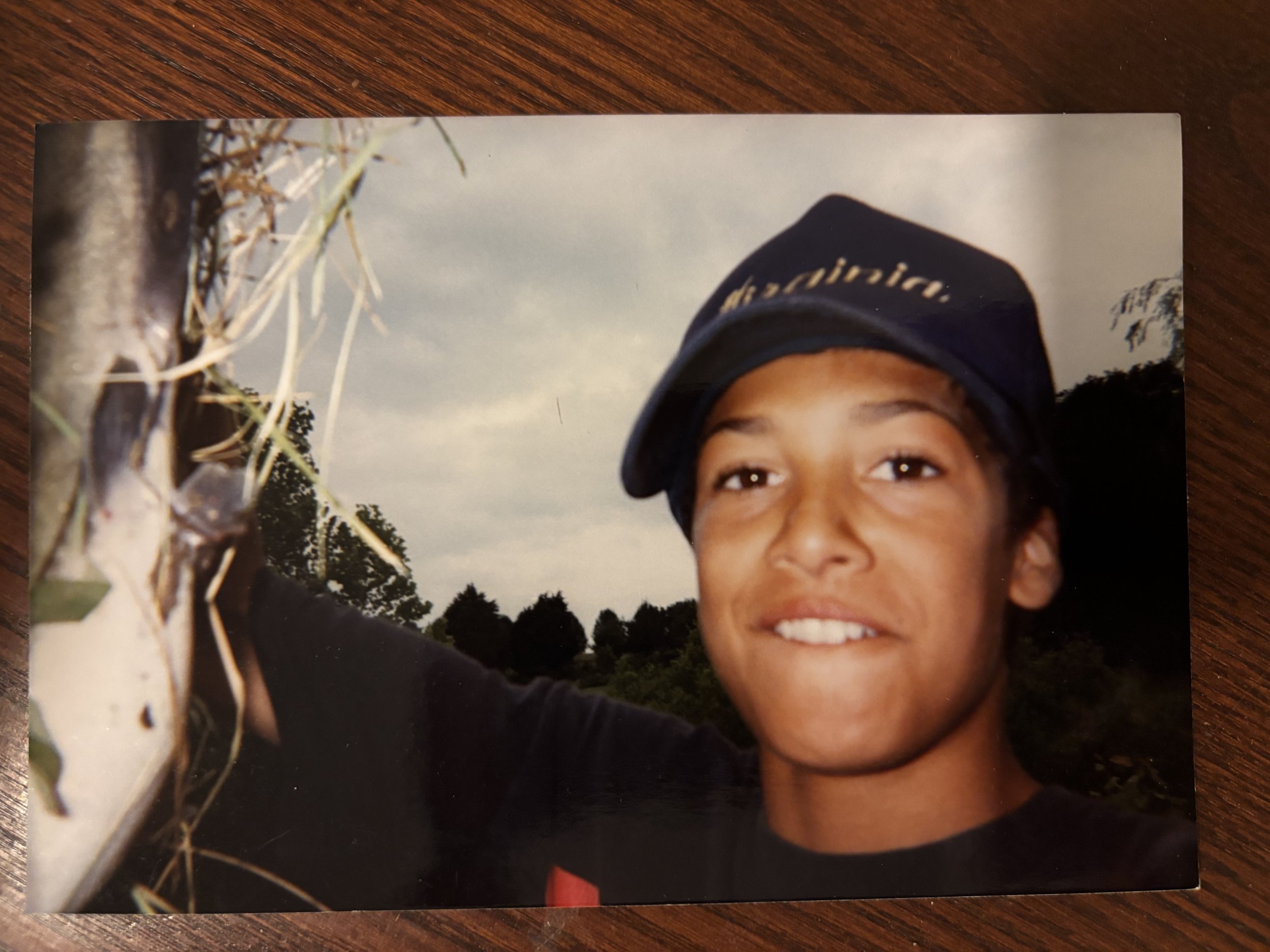 A young boy wearing a navy blue cap with the word 'dreamer' written on it, smiling outdoors with a cloudy sky and trees in the background.