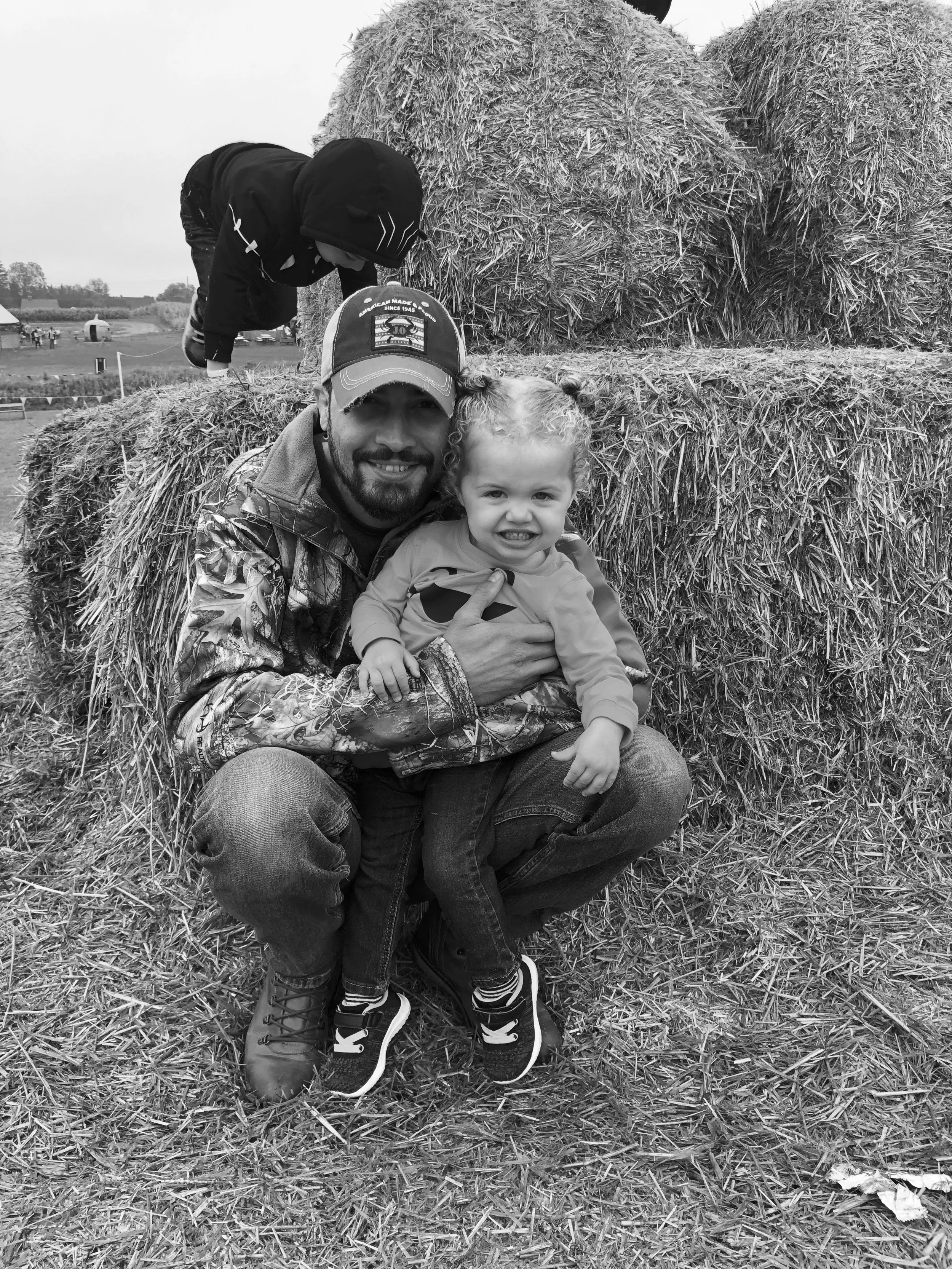 A man and a young girl with curly hair posing in front of stacked hay bales outdoors.