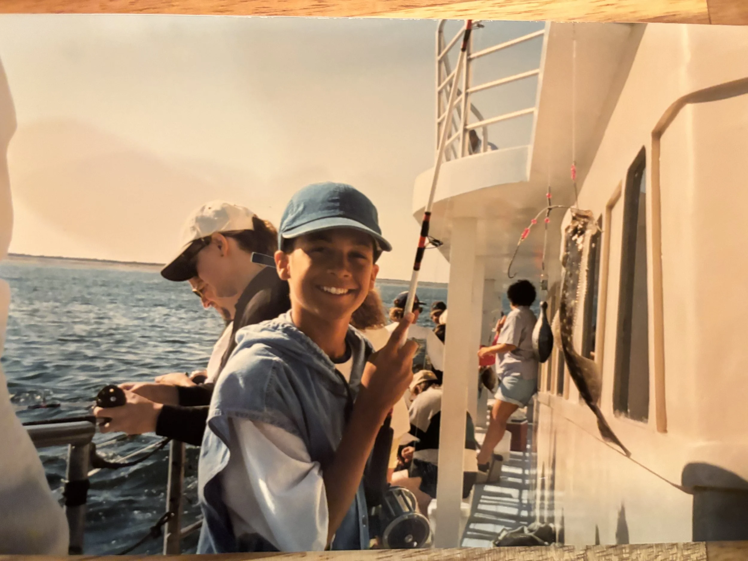 Smiling young man on a fishing boat holding a fishing rod with a fish caught on it, surrounded by other people fishing, with the ocean in the background.